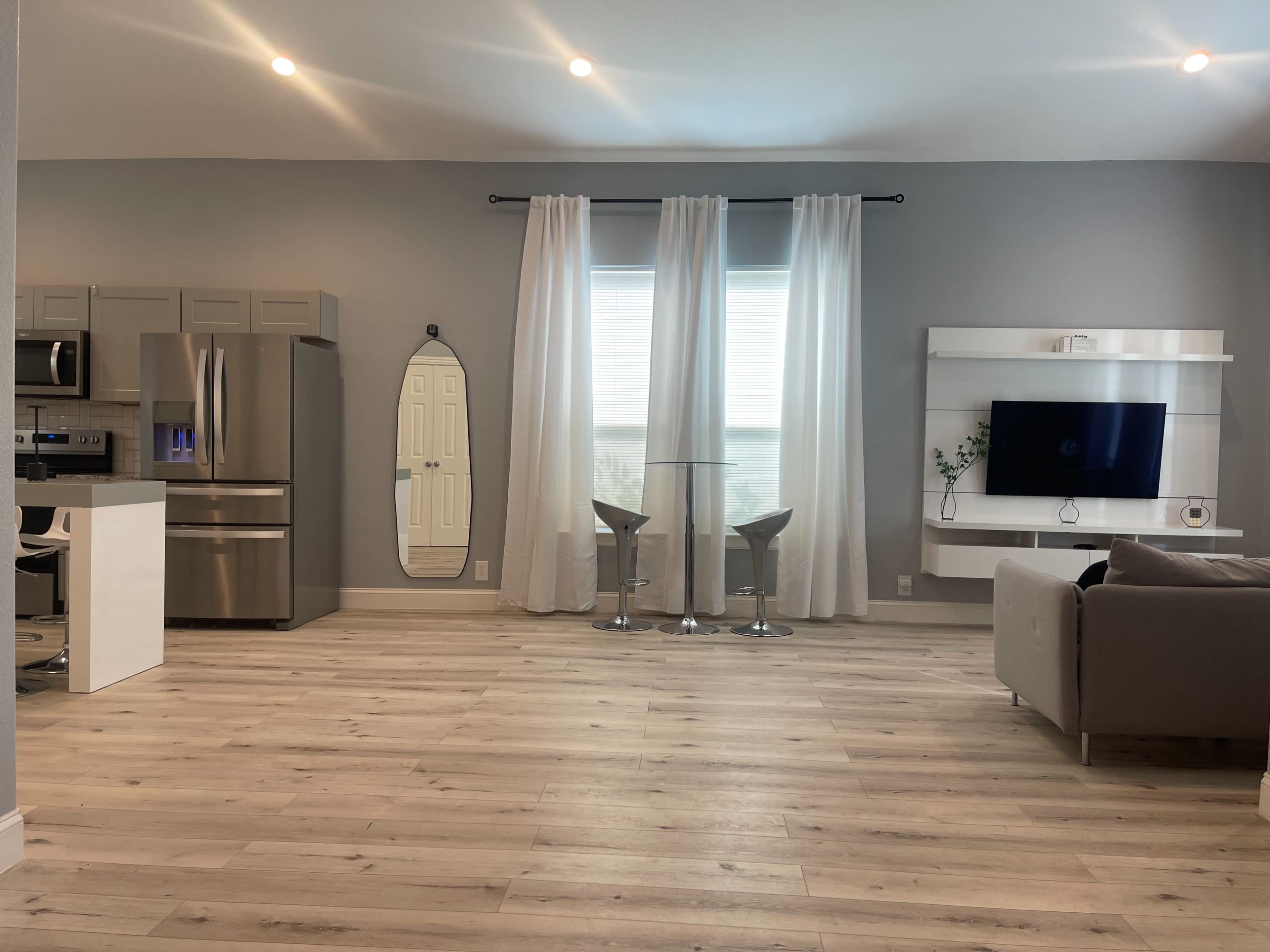 A modern living space features a kitchen area with stainless steel appliances, a large mirror, bar stools, and a television mounted on a white wall.