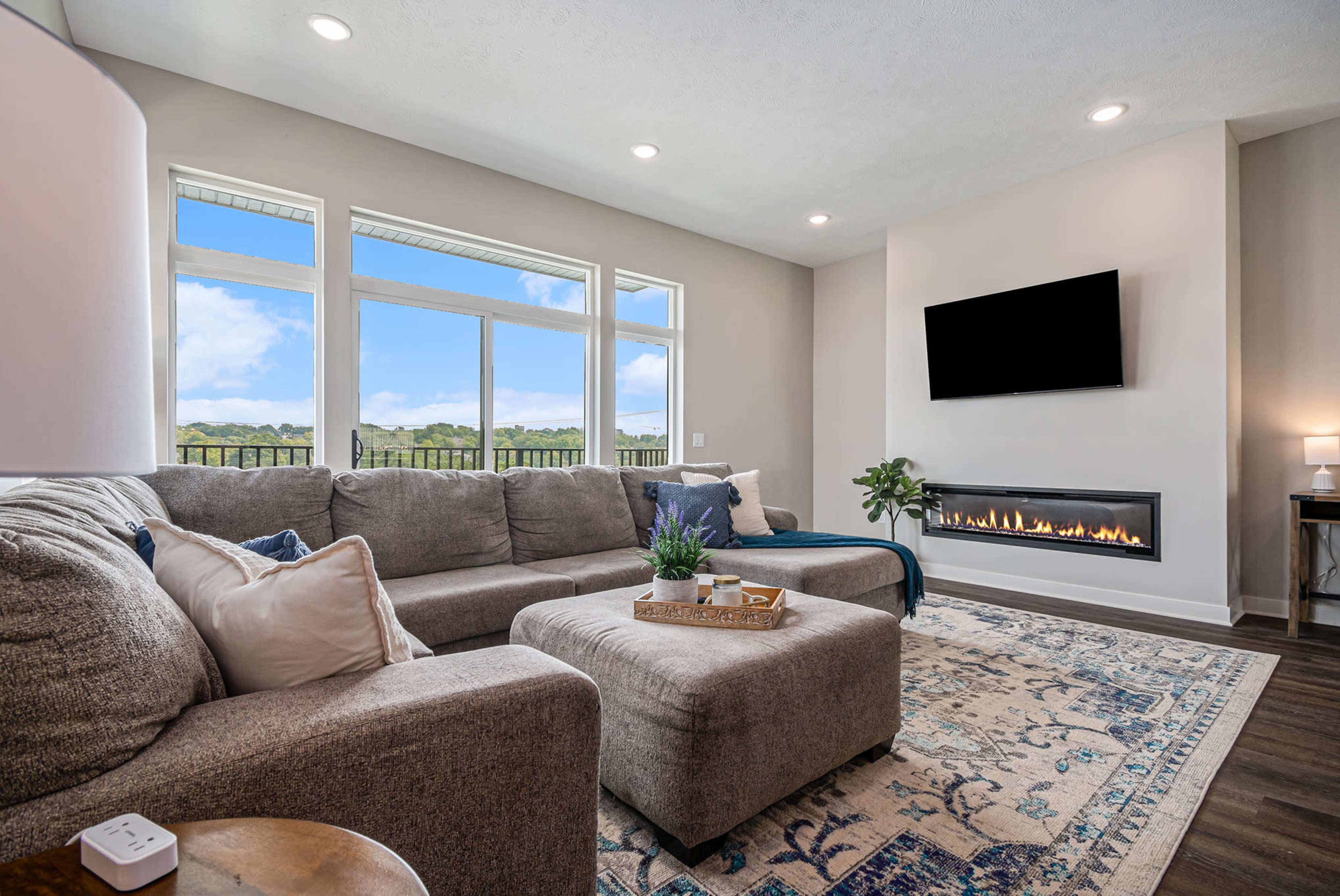A cozy living room features a sectional sofa, a wall-mounted TV, and a modern fireplace, with large windows offering a view of the outdoors.