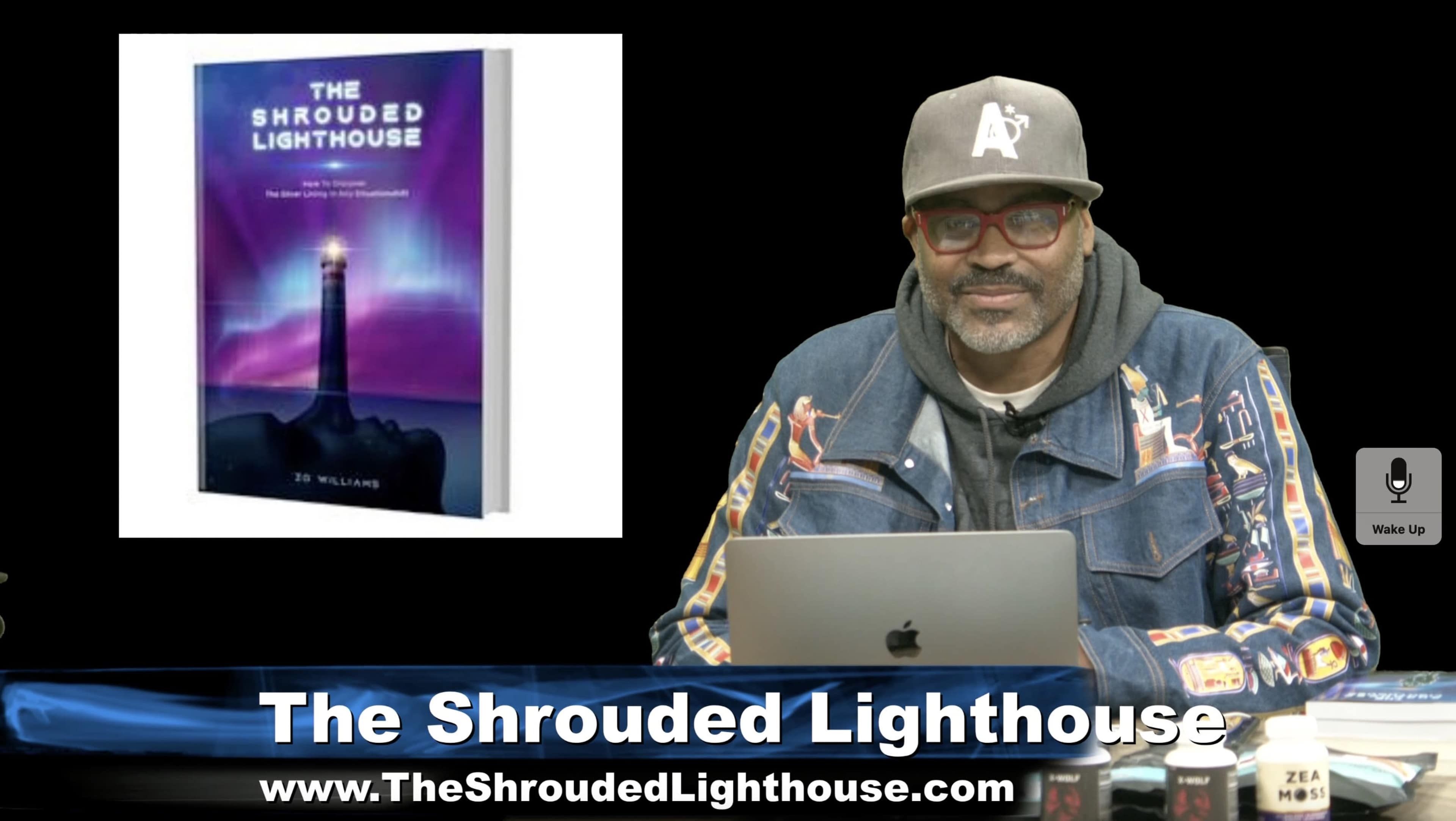 A man wearing glasses and a hat sits at a desk with a laptop, while a book titled "The Shrouded Lighthouse" is displayed beside him.