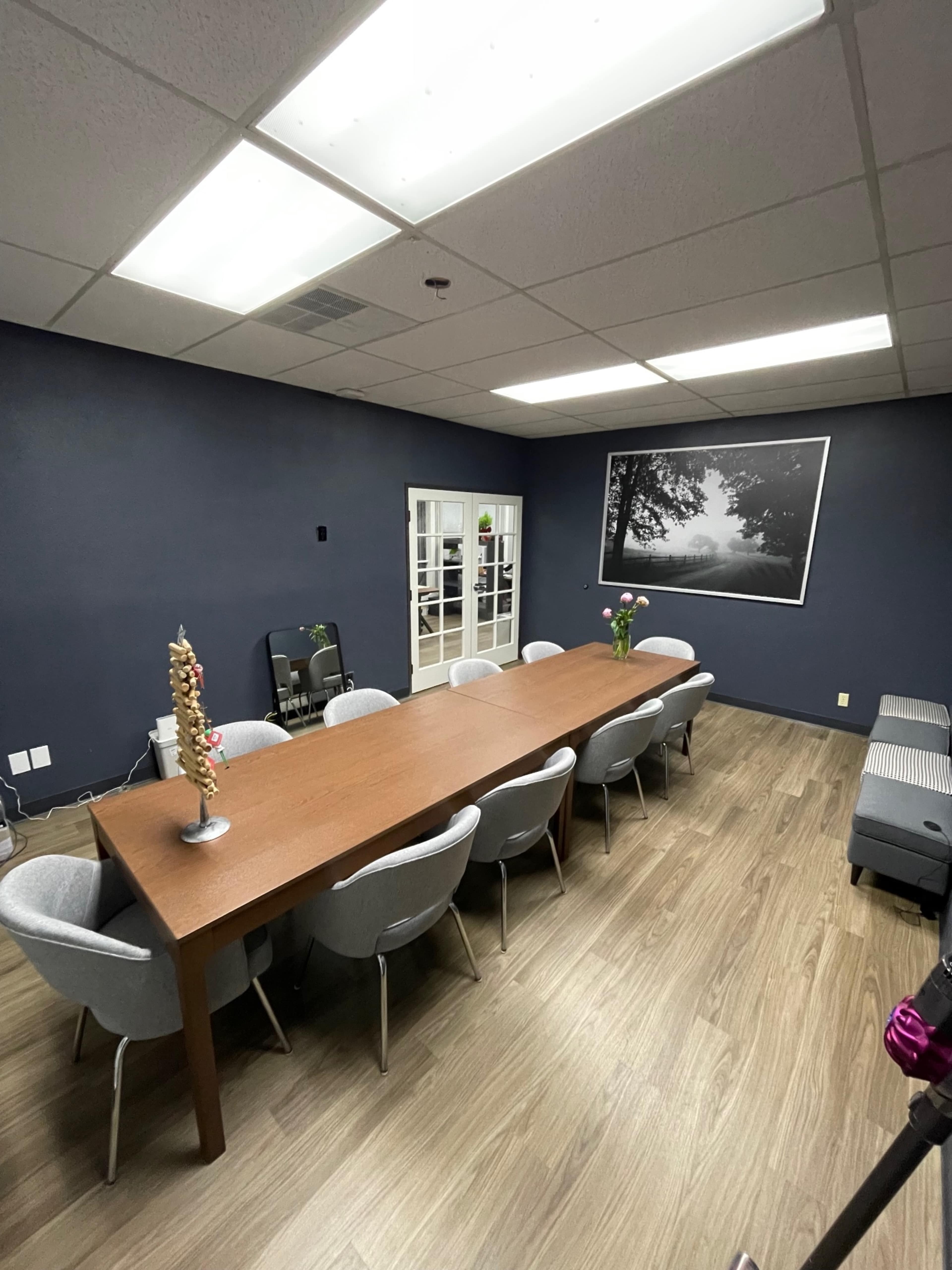 The image shows a conference room with a long wooden table surrounded by ten chairs, gray walls, and a large black-and-white photograph on one wall.