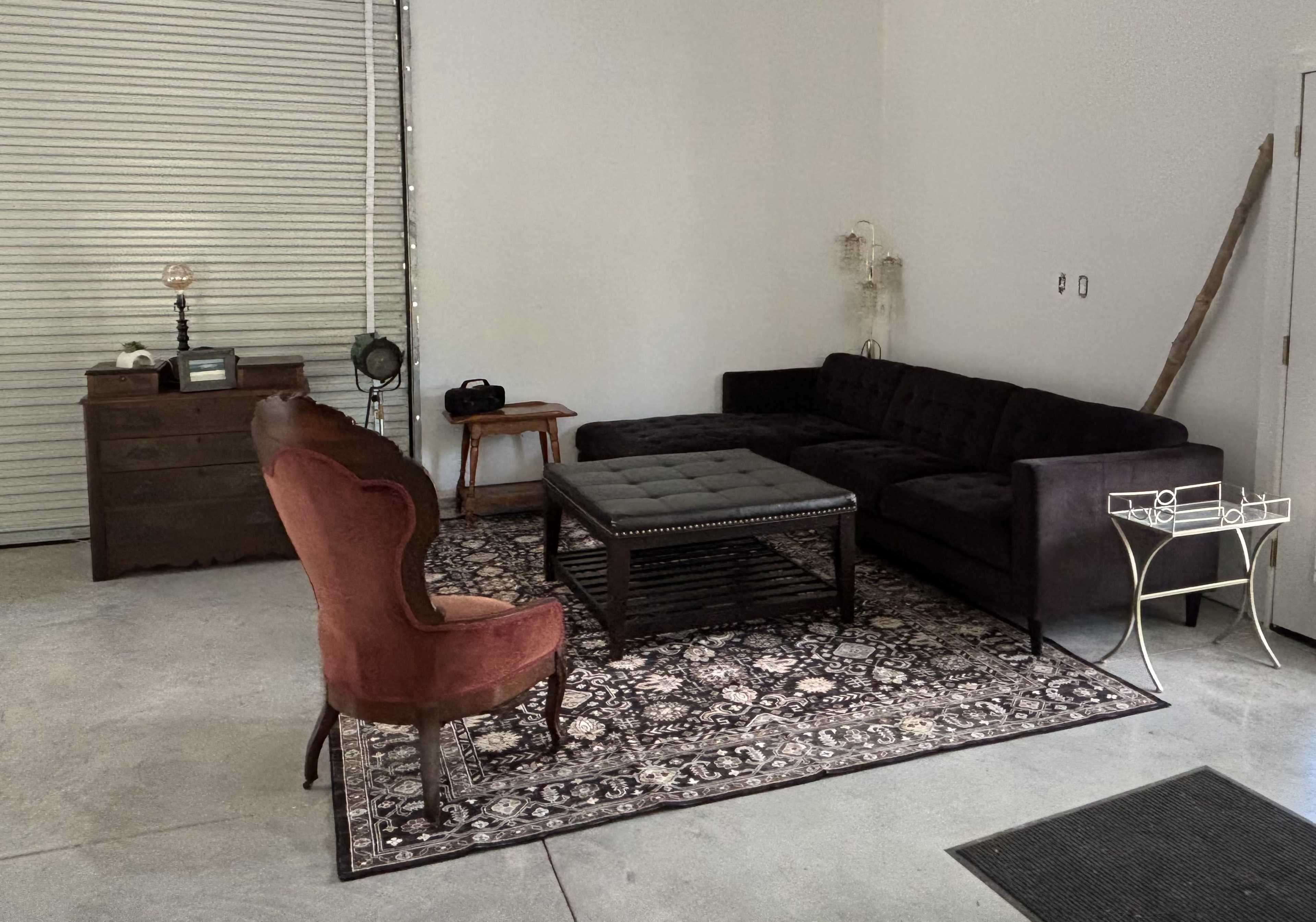 The image shows a minimalist room with a dark sectional sofa, an ornate rug, a vintage dresser, and an antique chair, all arranged around a coffee table.