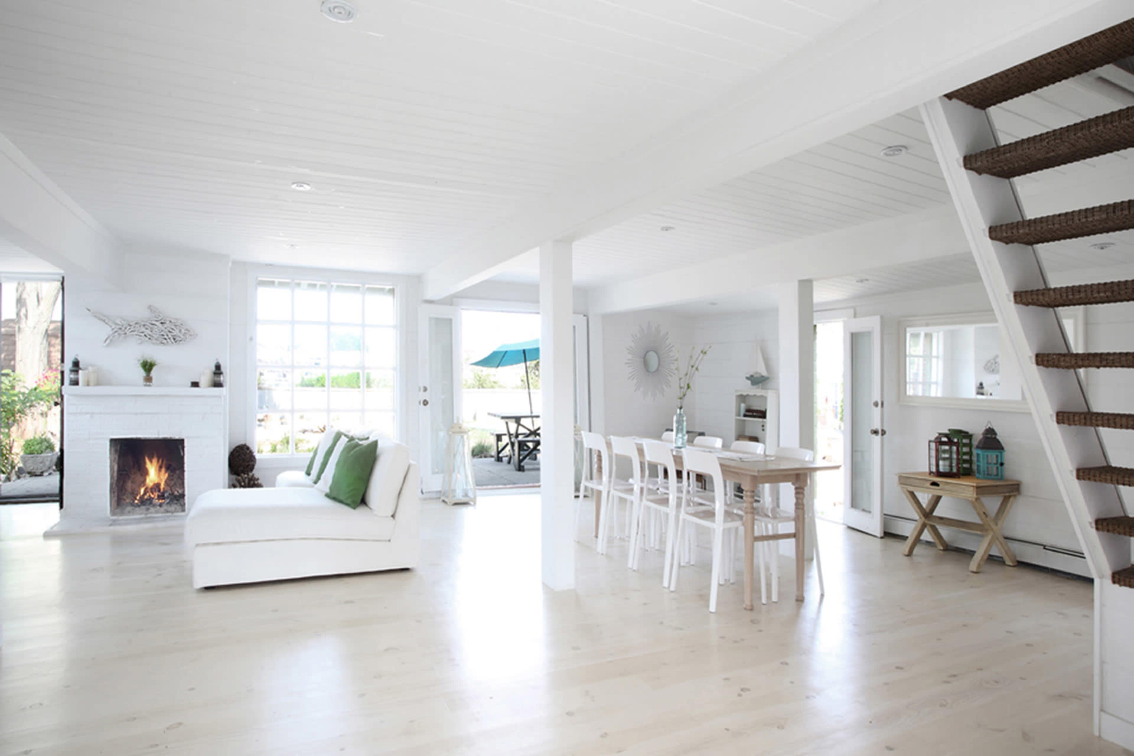 A bright, open-concept living space with a fireplace, dining table, and a staircase, featuring large windows that allow natural light to fill the room.