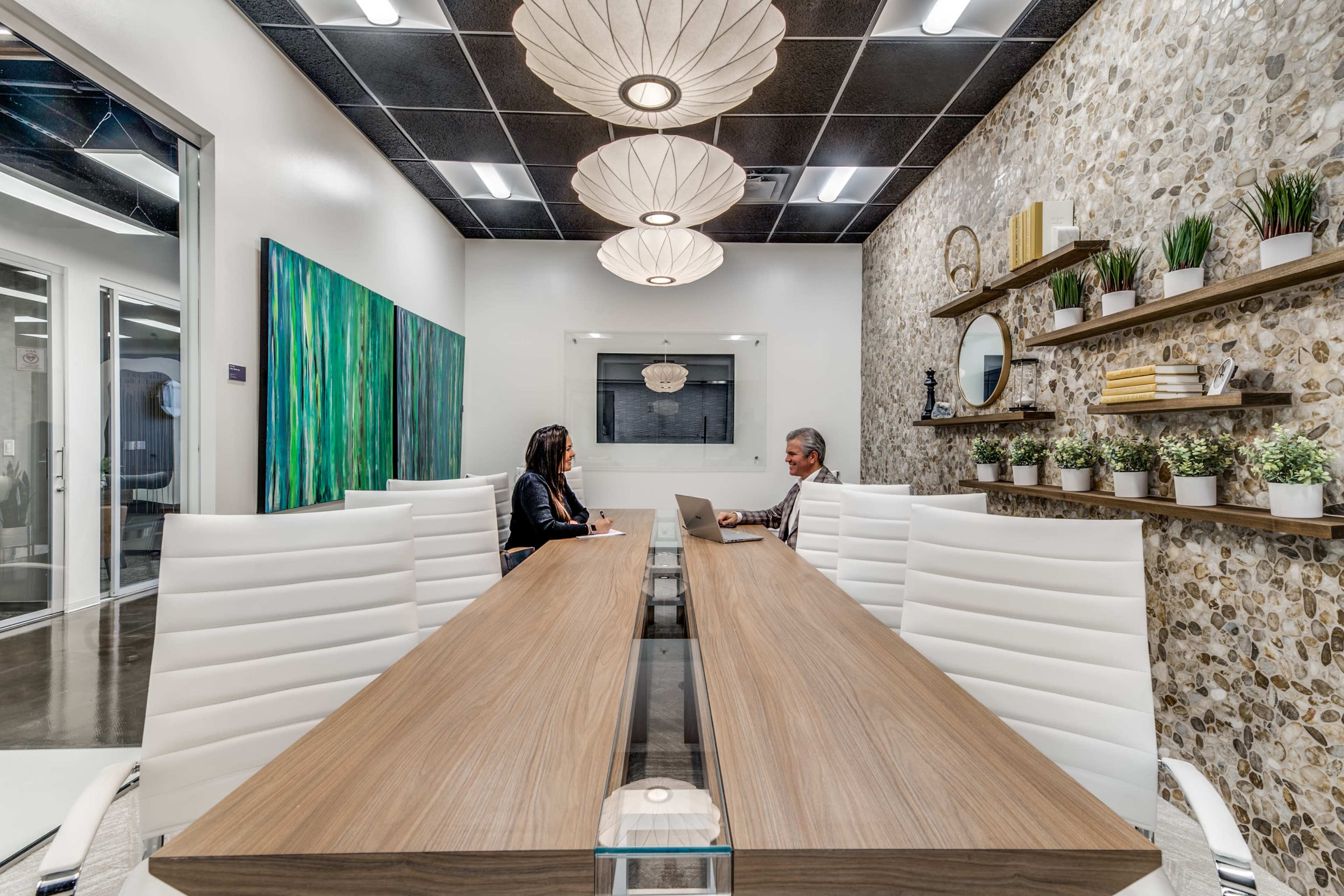 A modern conference room features a long wooden table, two people engaged in discussion, artwork on the walls, and decorative plants on shelves.