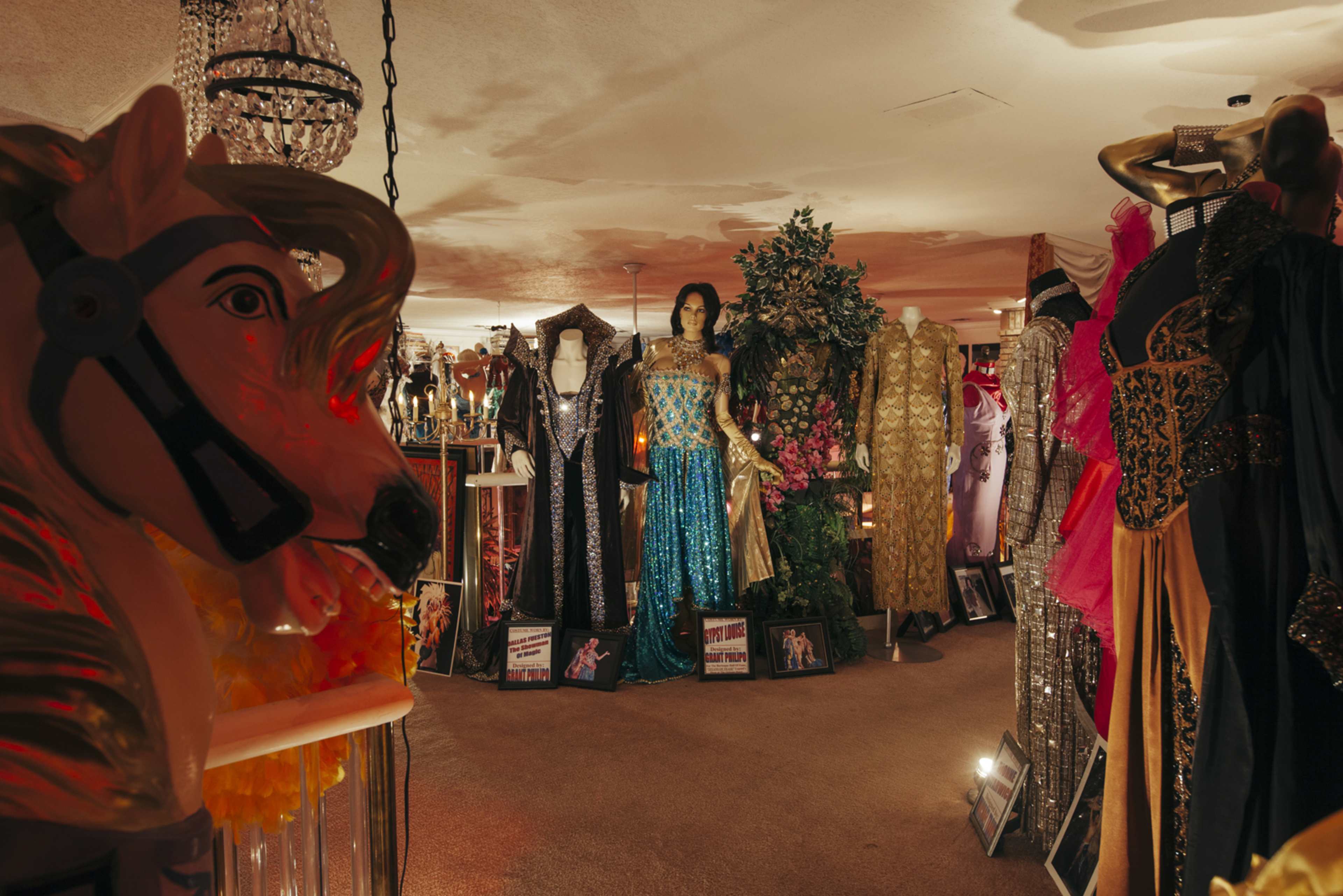 The image shows a vibrant boutique filled with a variety of evening gowns and costumes displayed among decorative elements, including a horse statue and chandeliers.