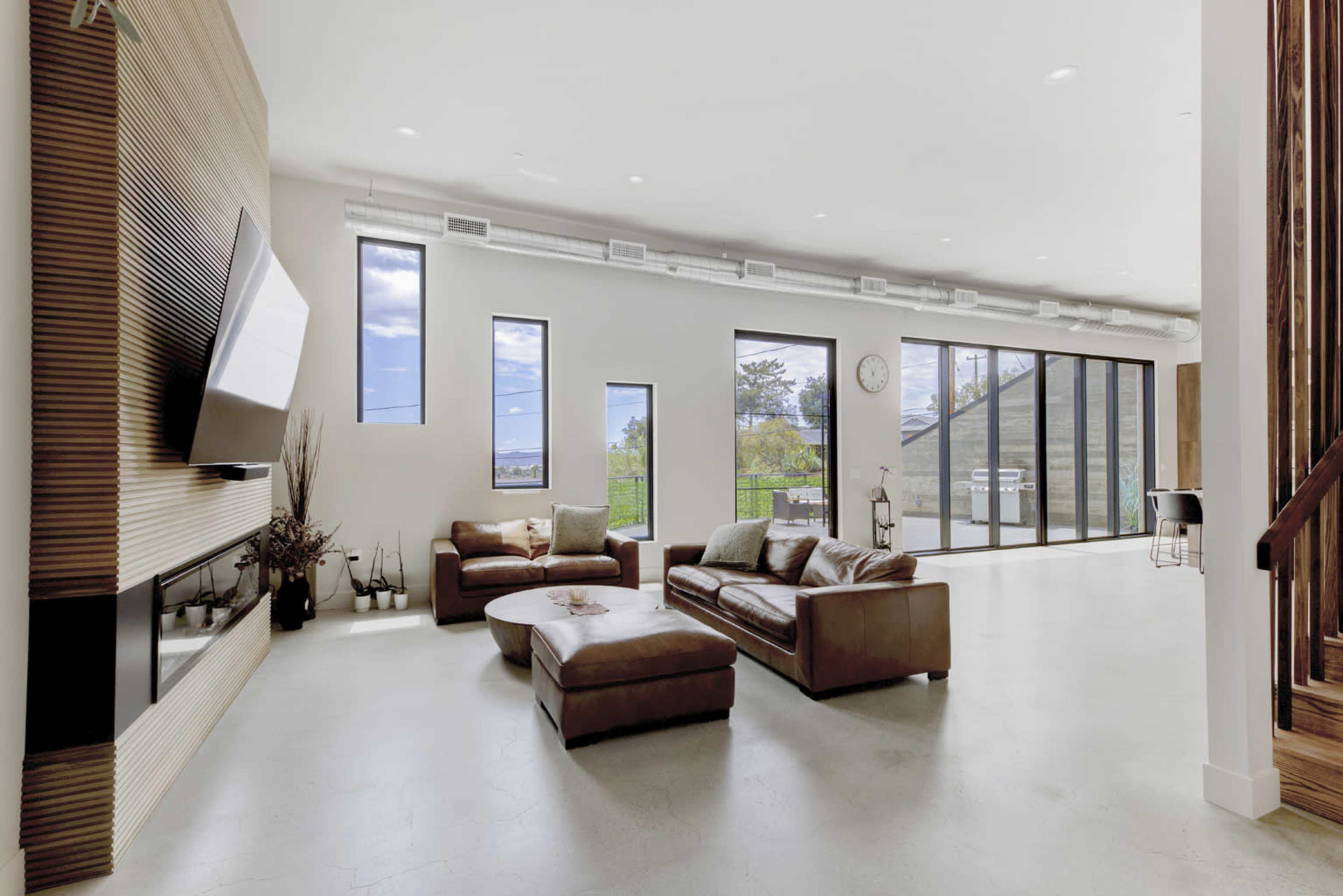 A spacious living room features large windows, a wall-mounted television, and modern furniture arranged around a coffee table.