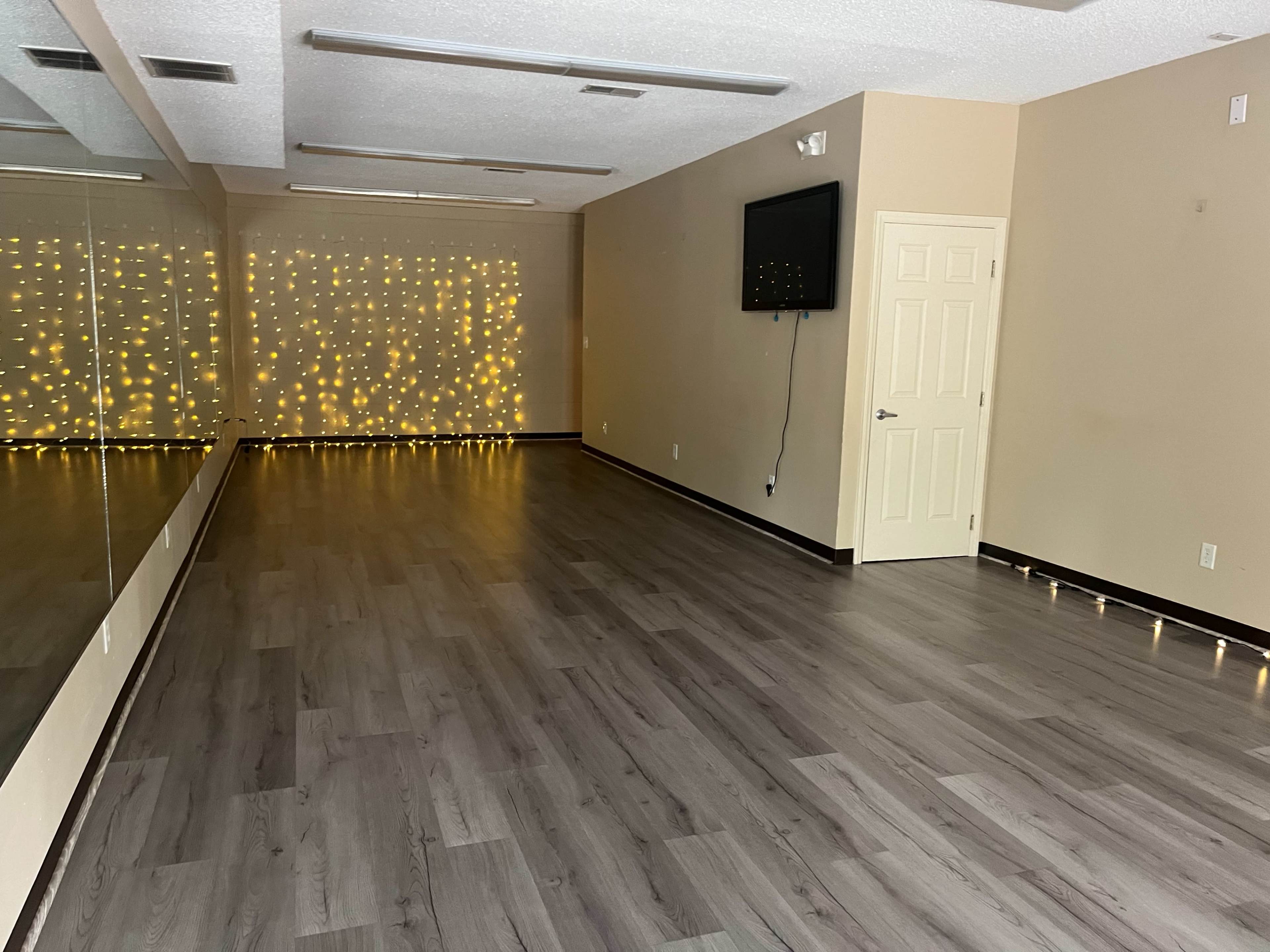 A spacious dance studio with wooden flooring, a wall of mirrors, and string lights in the background.