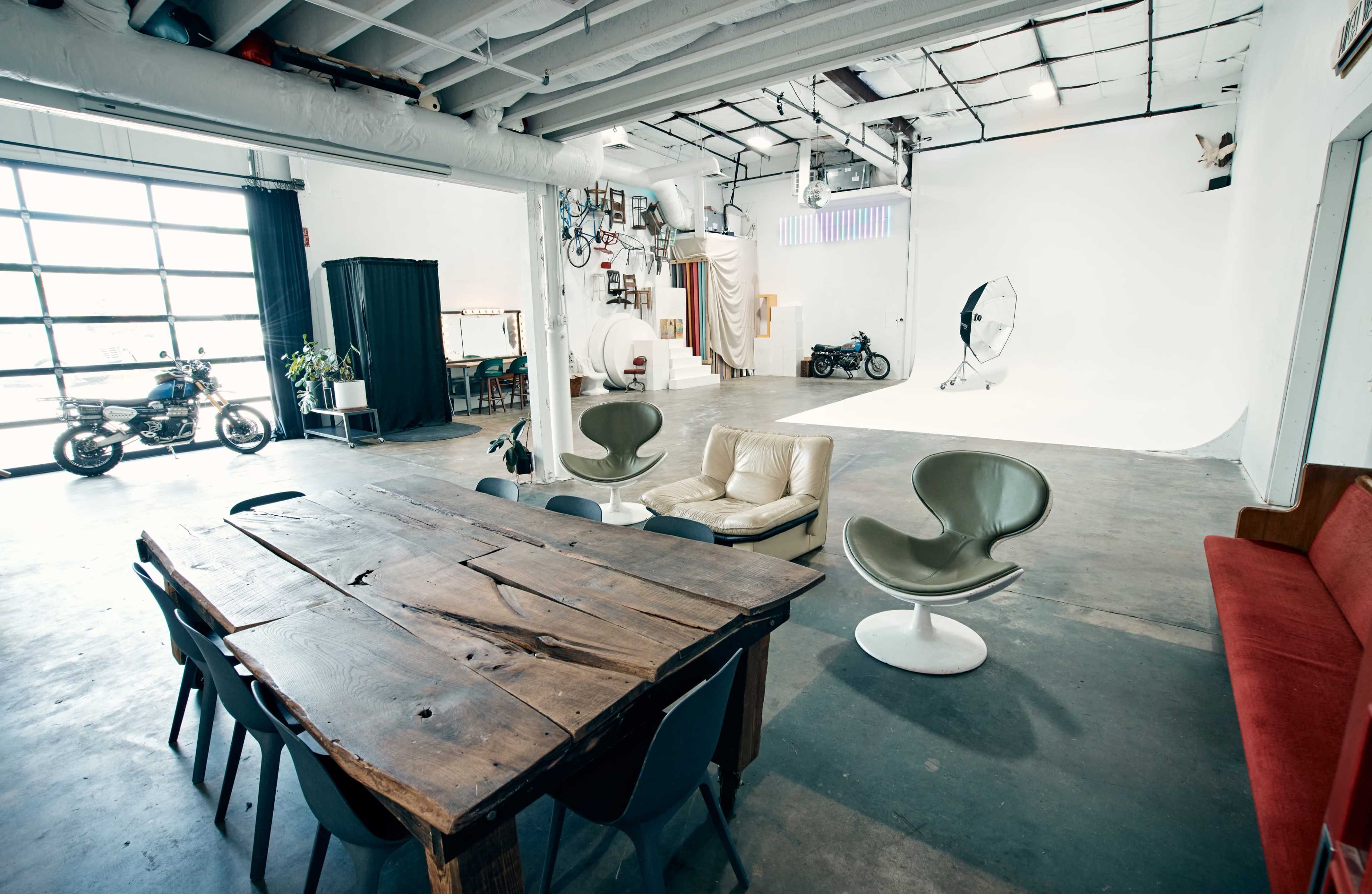 A spacious studio with a wooden dining table, multiple seating areas, and a large white backdrop for photography, alongside a motorcycle and decor on the walls.