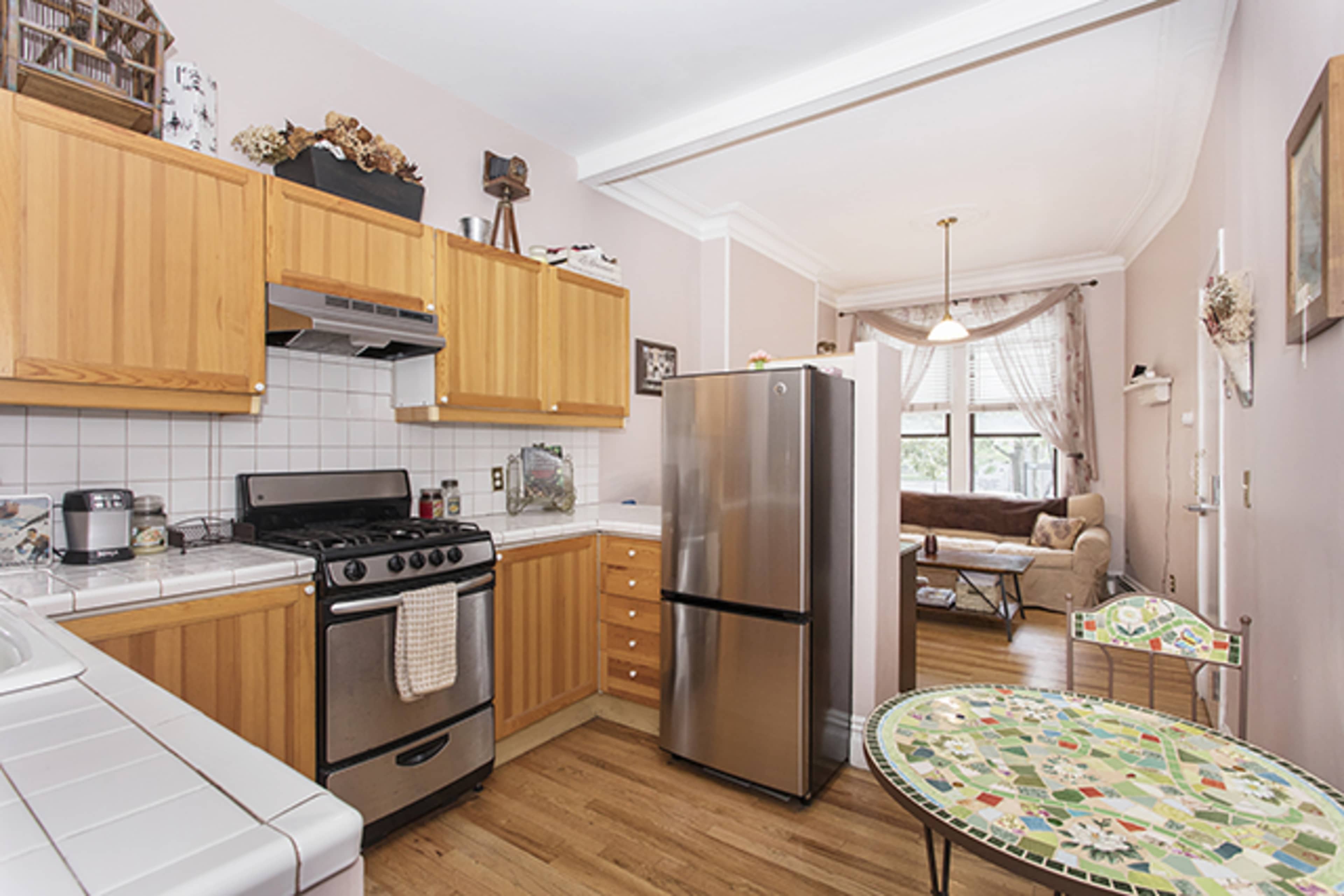 The image shows a kitchen with wooden cabinetry, a stainless steel refrigerator, a gas stove, and a small mosaic-topped table, leading into a living area with a couch.