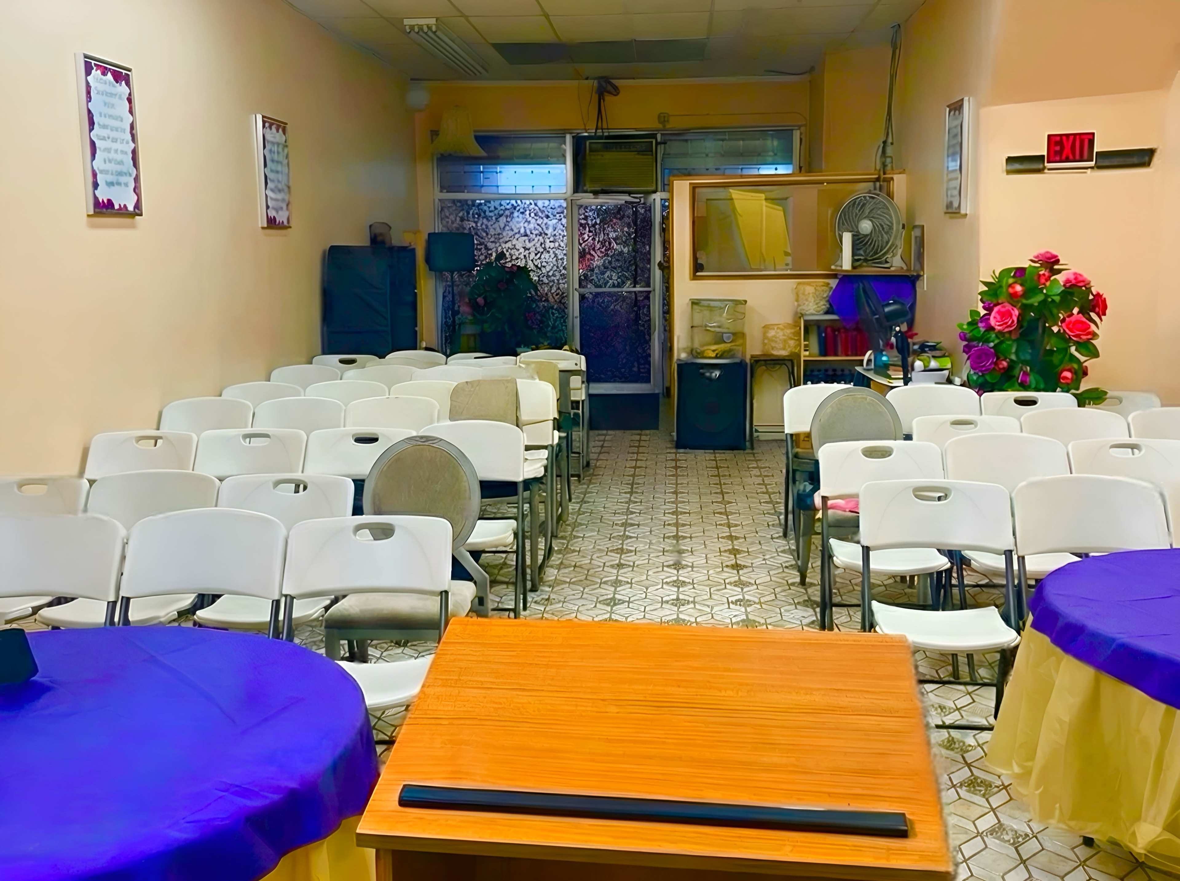 The image shows a small indoor room with rows of white chairs facing a wooden table, flanked by two round tables covered with purple tablecloths, and a decorative arrangement of flowers in the corner.