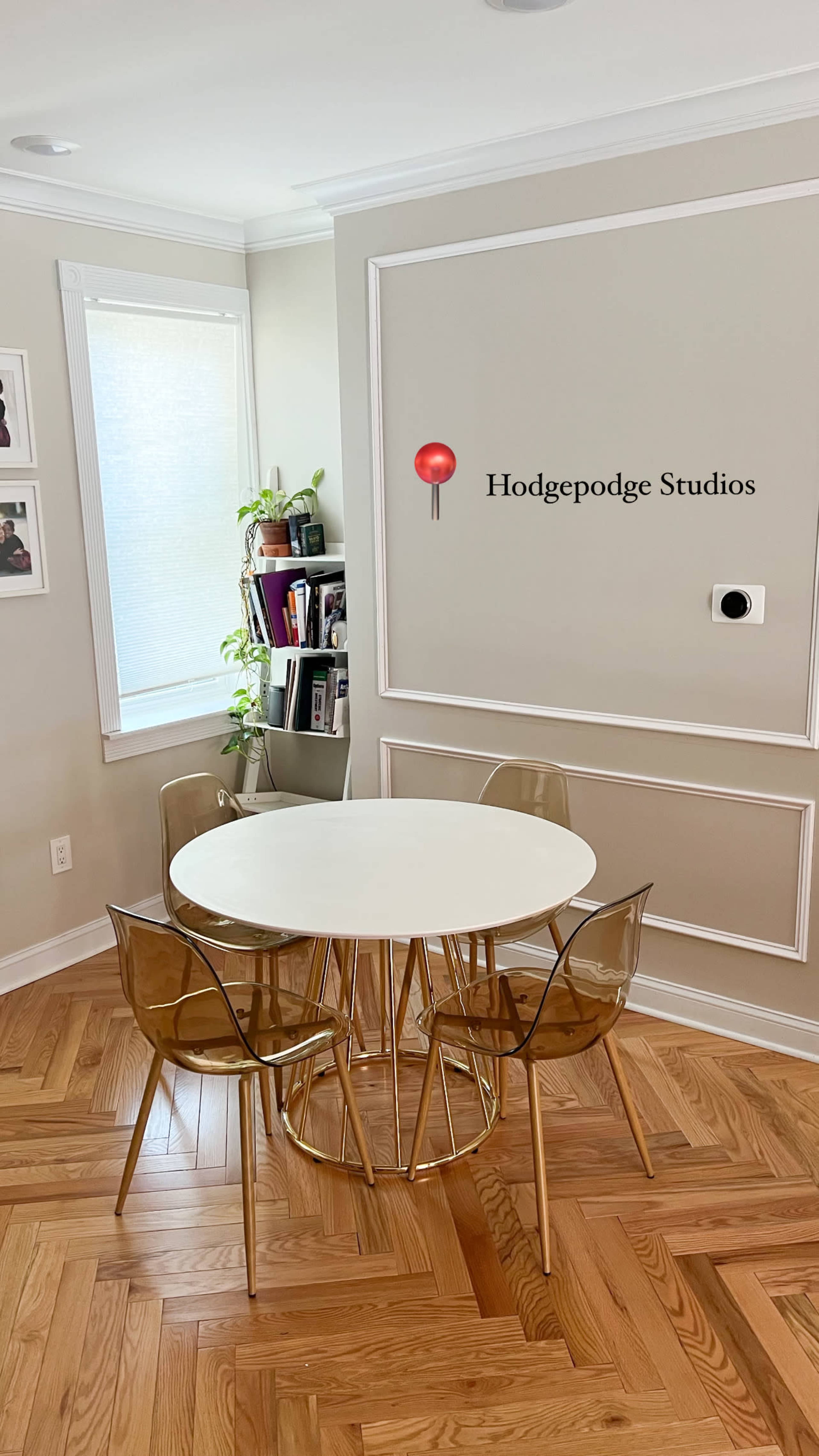 A round white table is surrounded by clear acrylic chairs in a well-lit room featuring a plant and shelves on one side.