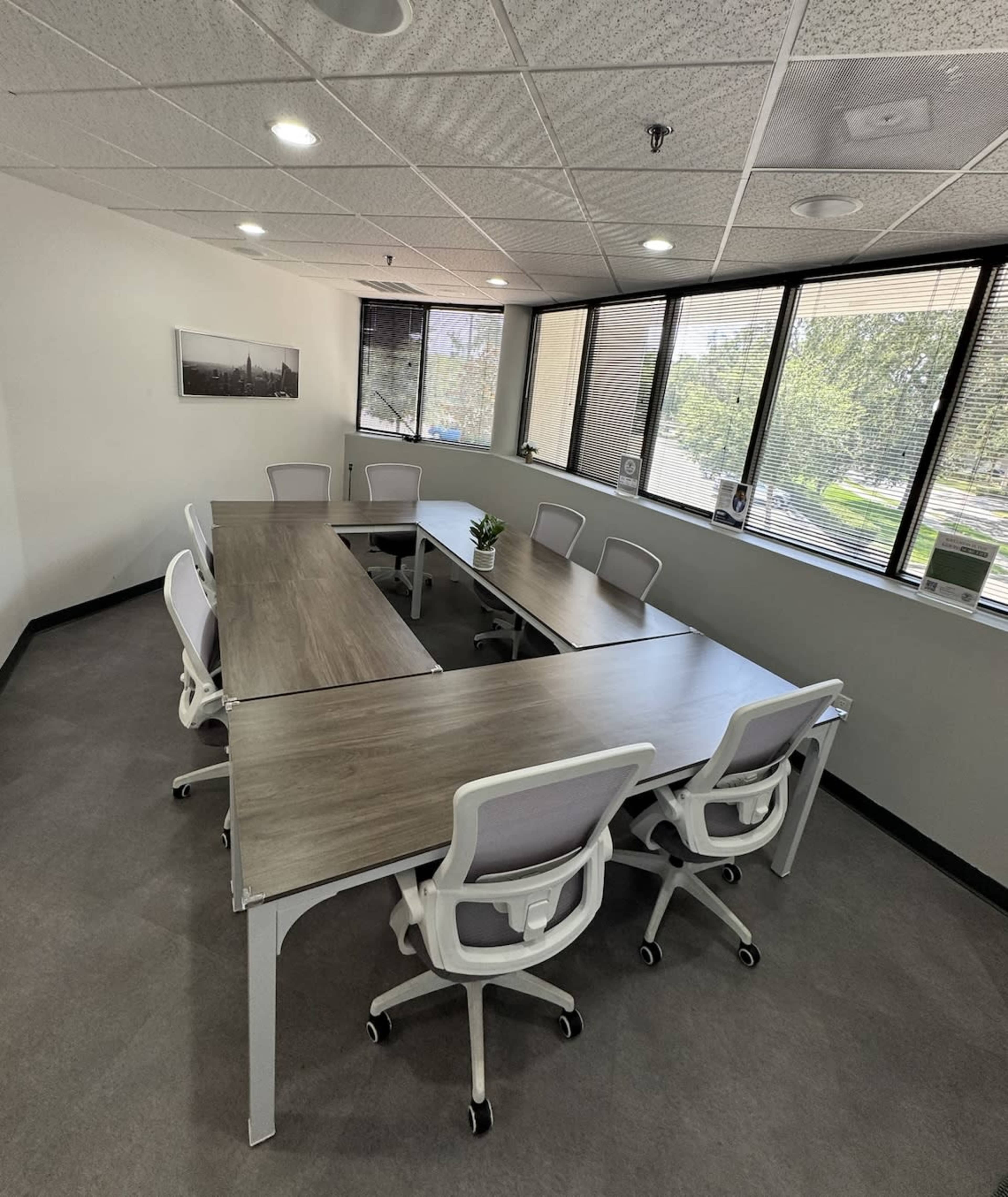 The image shows a modern conference room with two connected tables surrounded by eight ergonomic chairs and large windows letting in natural light.