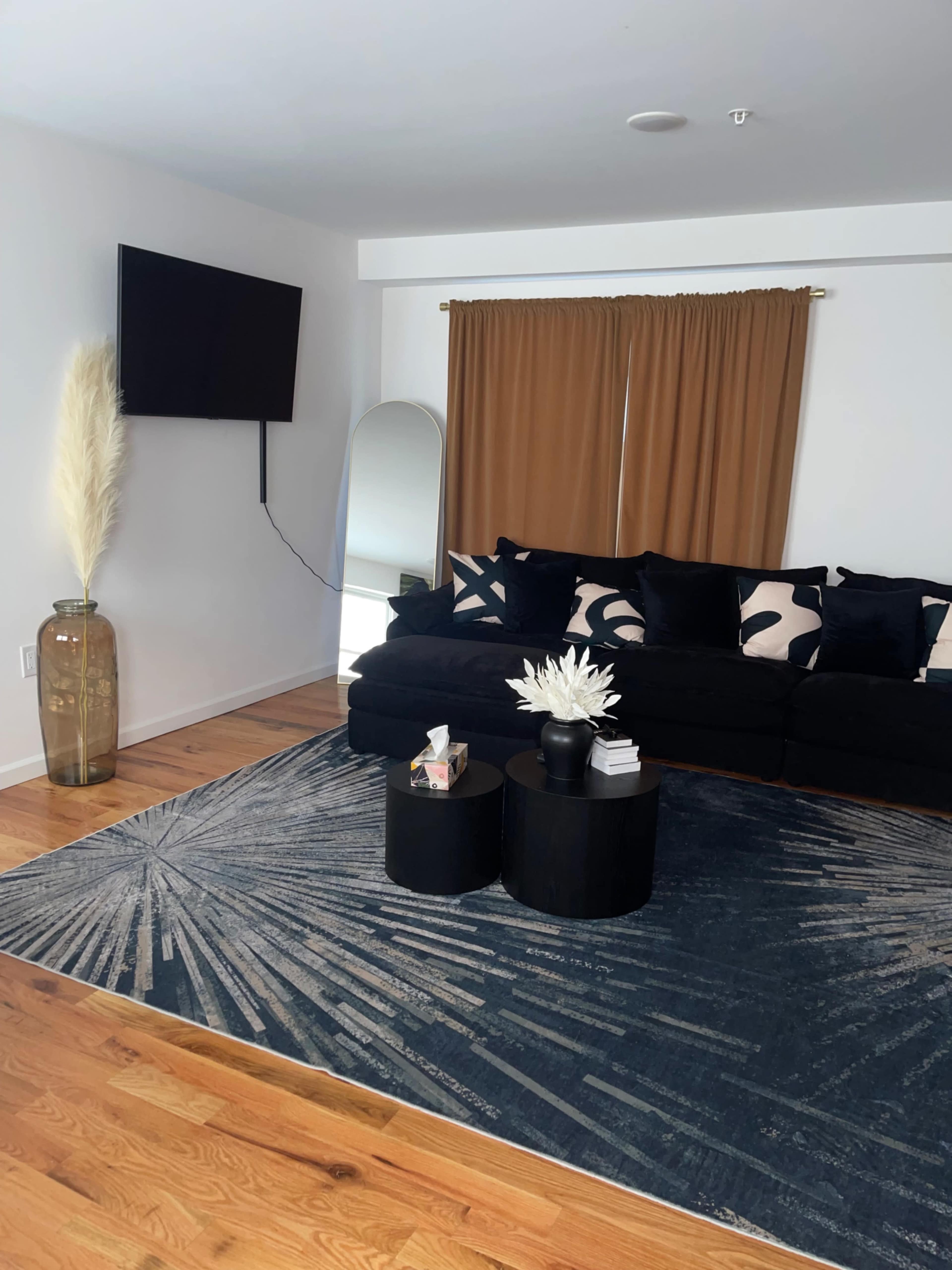 The living room features a black sofa with decorative pillows, a round black coffee table, a large area rug with a starburst pattern, a tall mirror, a flat-screen TV mounted on the wall, and curtains in a warm shade.