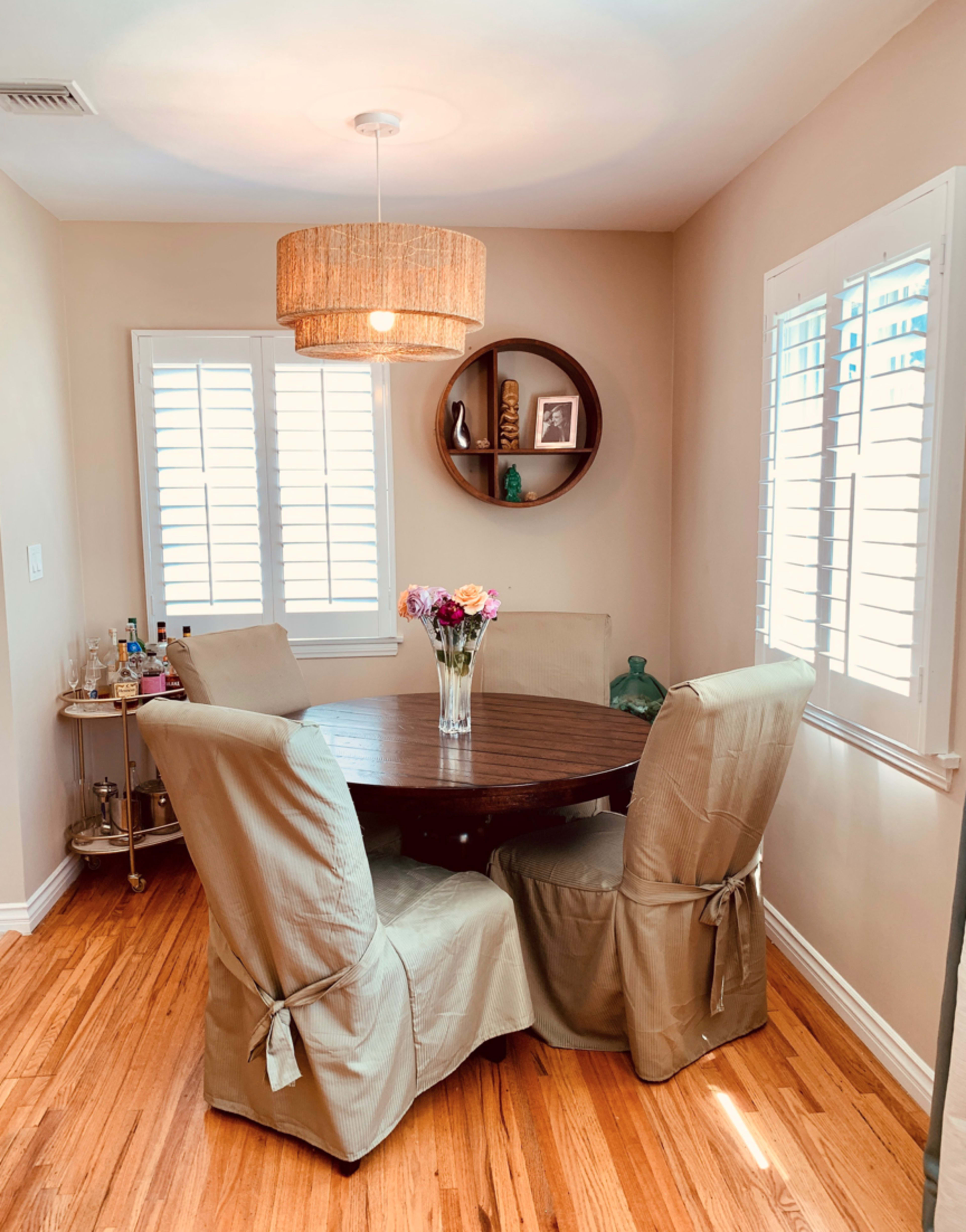 A round wooden dining table with four beige upholstered chairs is set in a bright room featuring a decorative light fixture and a wall shelf.
