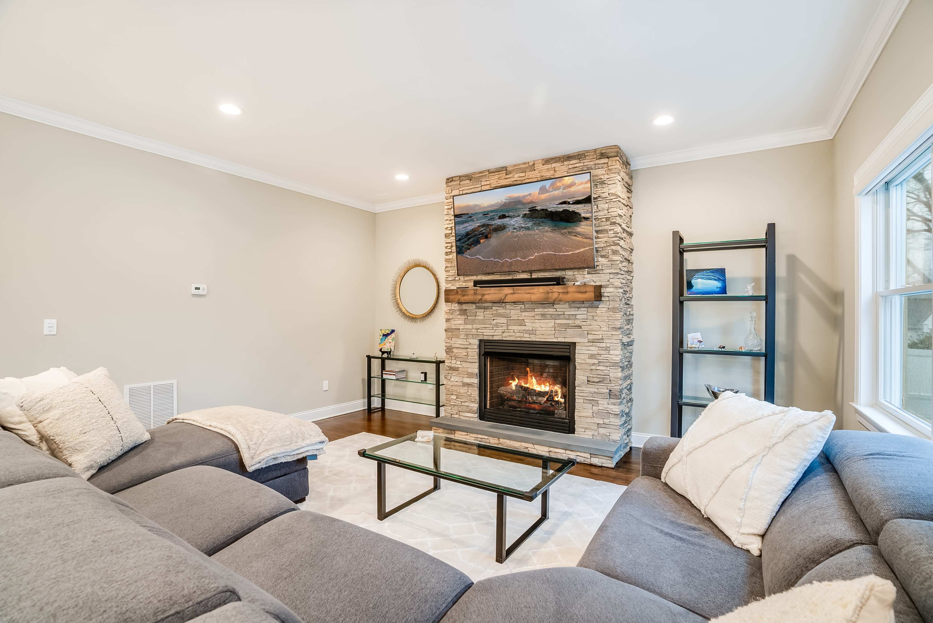 A modern living room features a gray sectional sofa, a glass coffee table, a stone fireplace with a television mounted above, and a window with natural light.