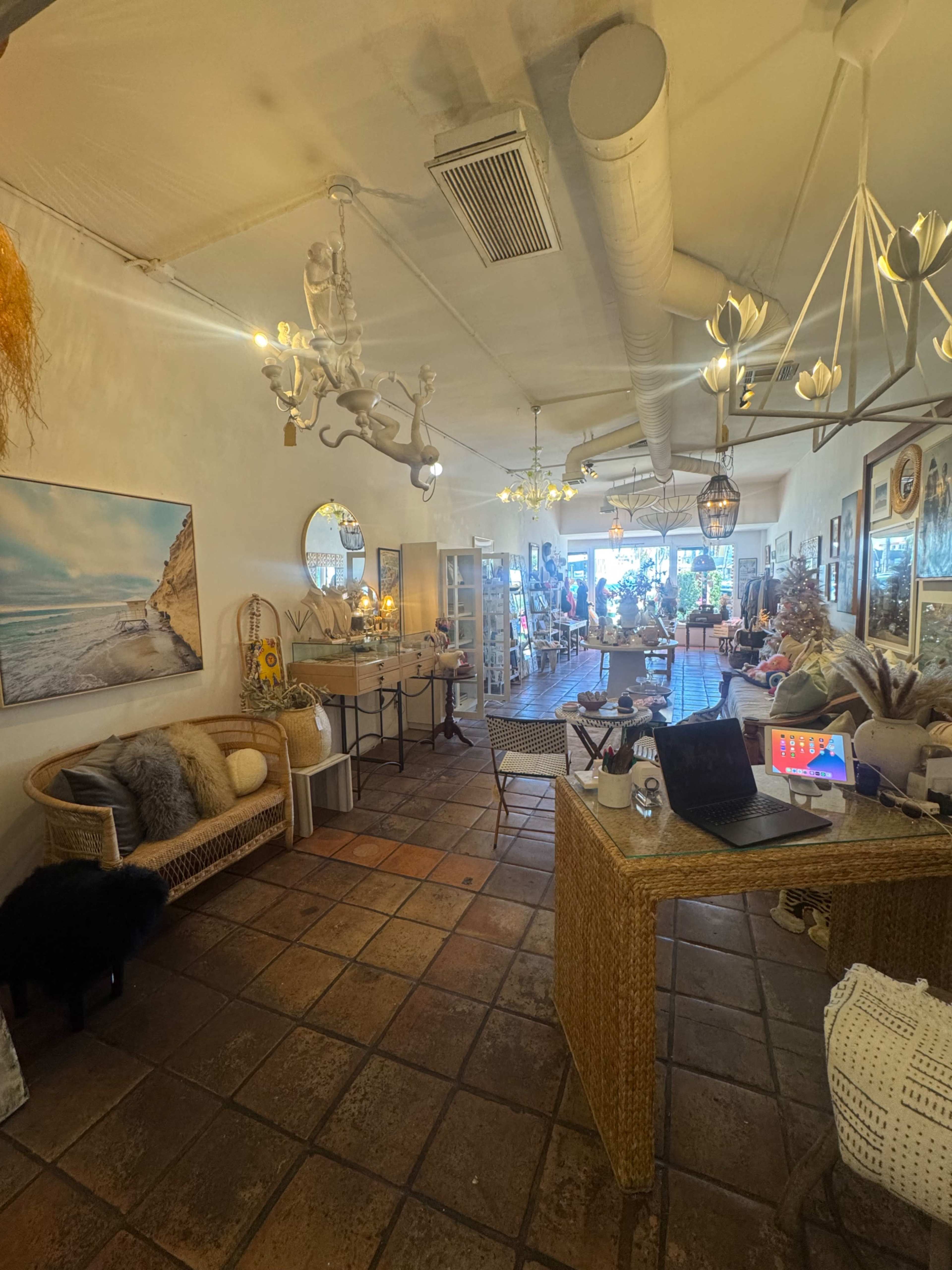 The image shows a cozy, well-decorated interior of a boutique featuring a variety of furniture, lighting fixtures, and decorative items, with natural light coming from large windows.