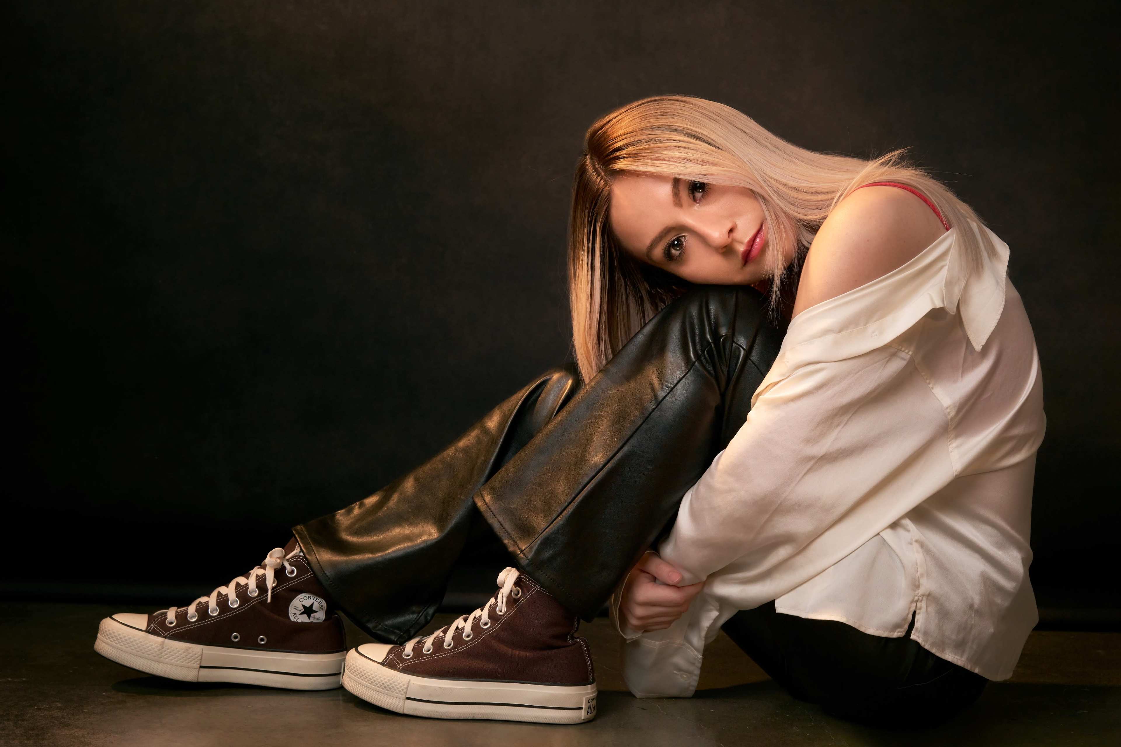A person sits on the floor with their knees drawn up, wearing a loose white shirt and black leather pants, while looking directly at the camera.