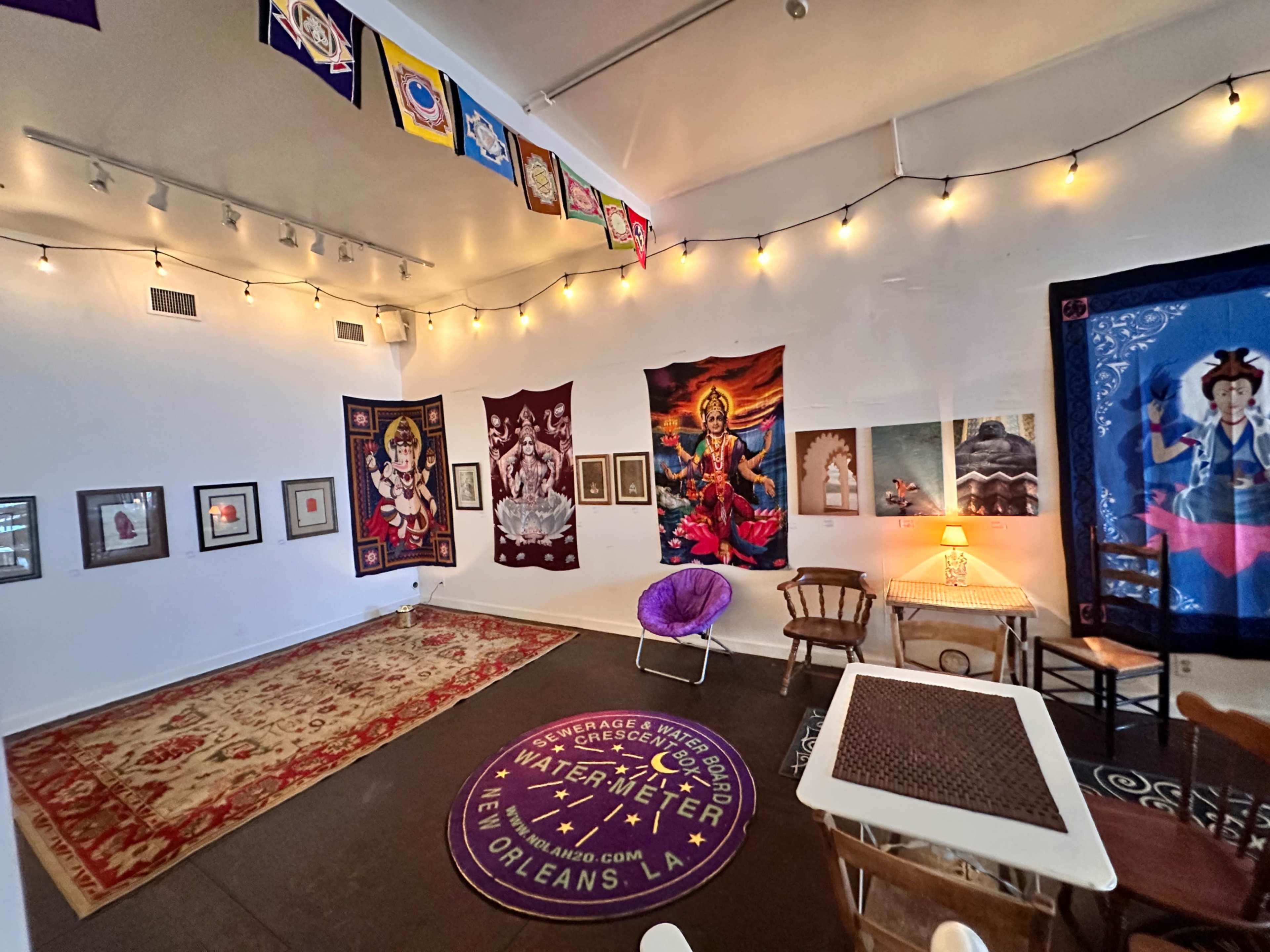The image shows a brightly lit room adorned with colorful tapestries and framed artwork, featuring a mix of decorative furniture and a patterned carpet.