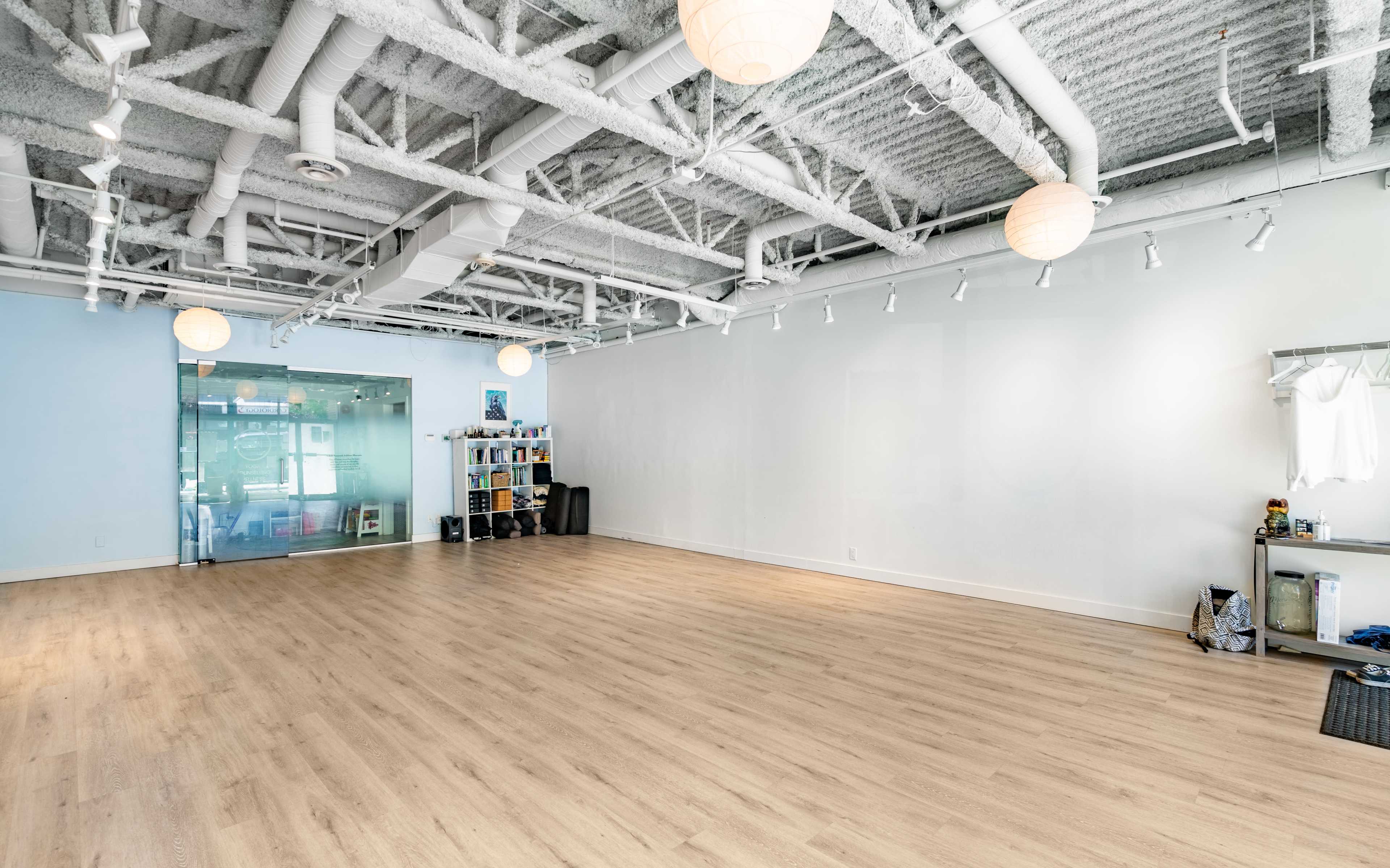 A spacious, well-lit studio with wooden flooring and a minimalistic design, featuring hanging light fixtures and shelves along one wall.