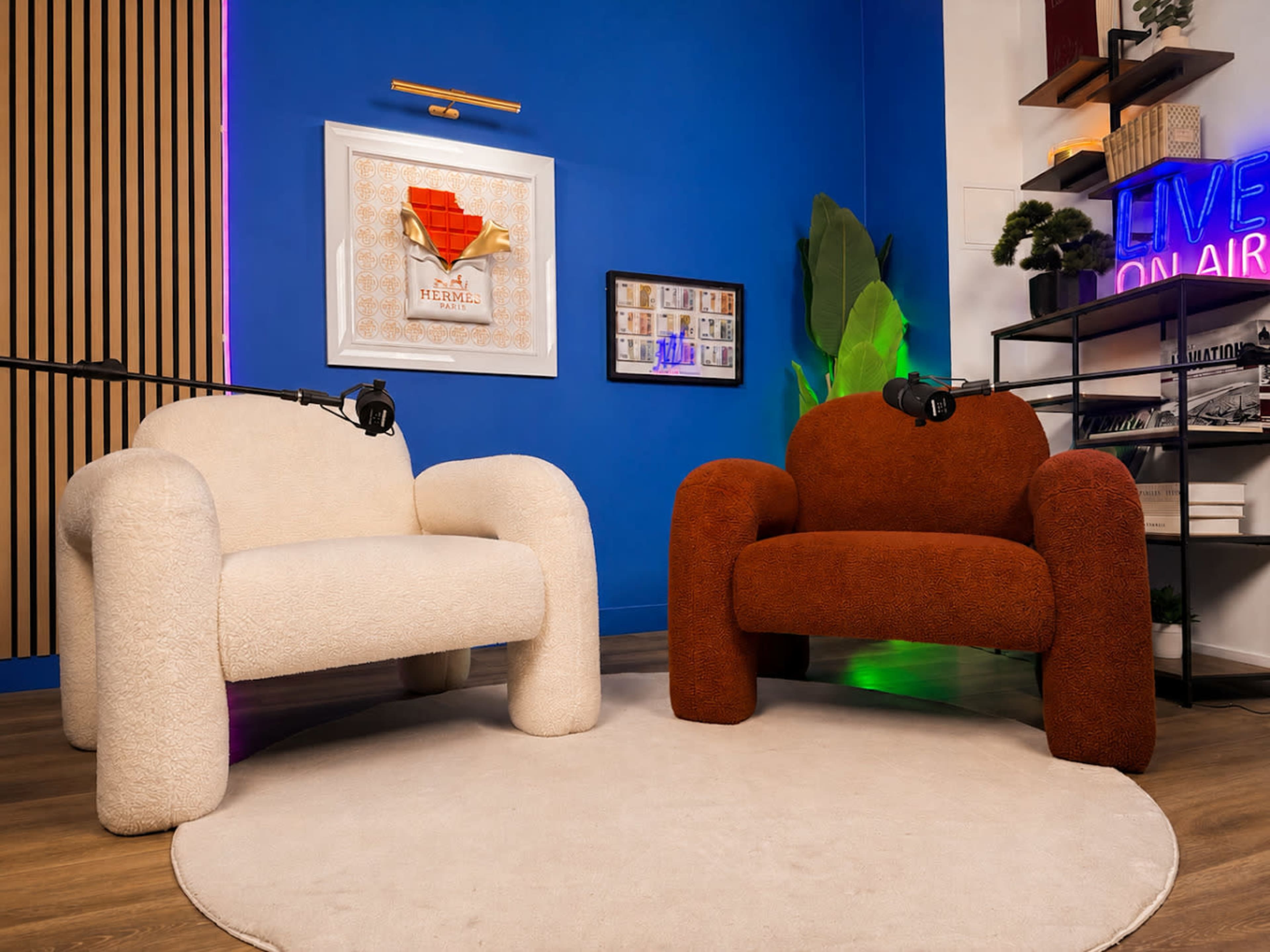 The image features two uniquely shaped armchairs, one cream-colored and the other rust-colored, positioned on a circular rug against a blue wall with decorative elements.