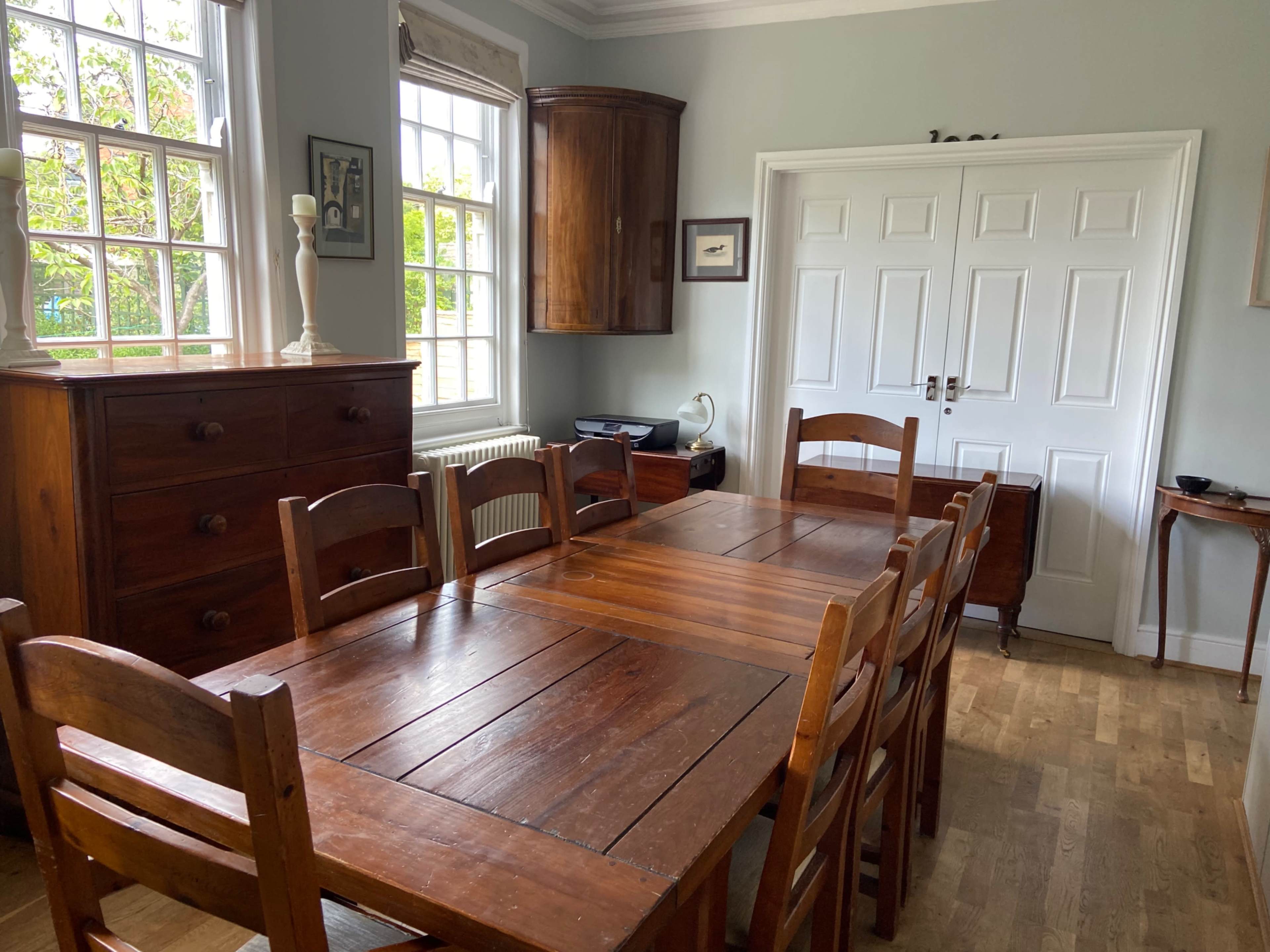A wooden dining table surrounded by chairs is positioned in a bright room with windows and wooden furniture.