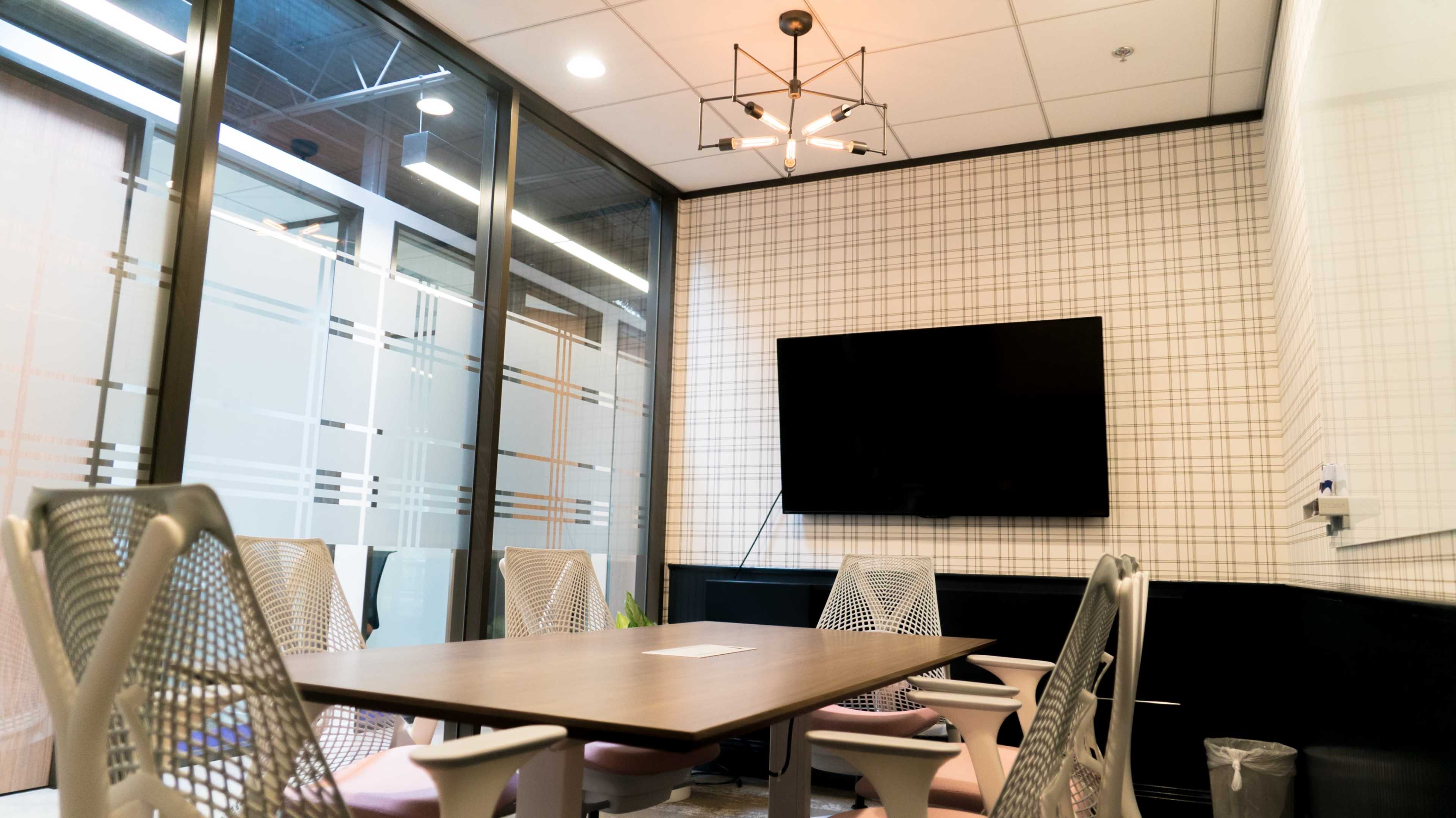 A modern conference room features a large table surrounded by ergonomic chairs, a wall-mounted television, and a grid-patterned backdrop.