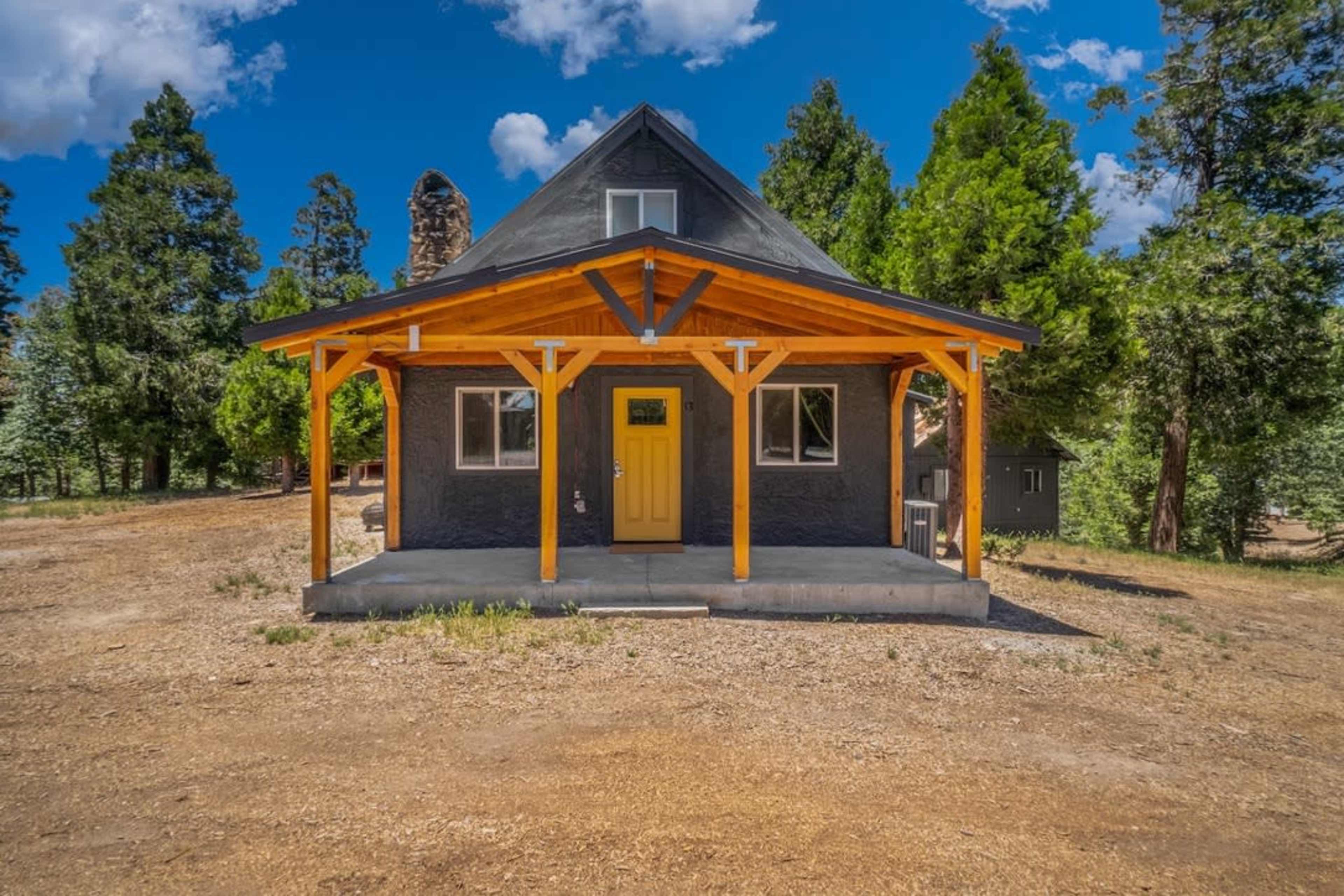 A two-story cabin with a yellow door and a wooden porch is situated on a gravel lot surrounded by trees.