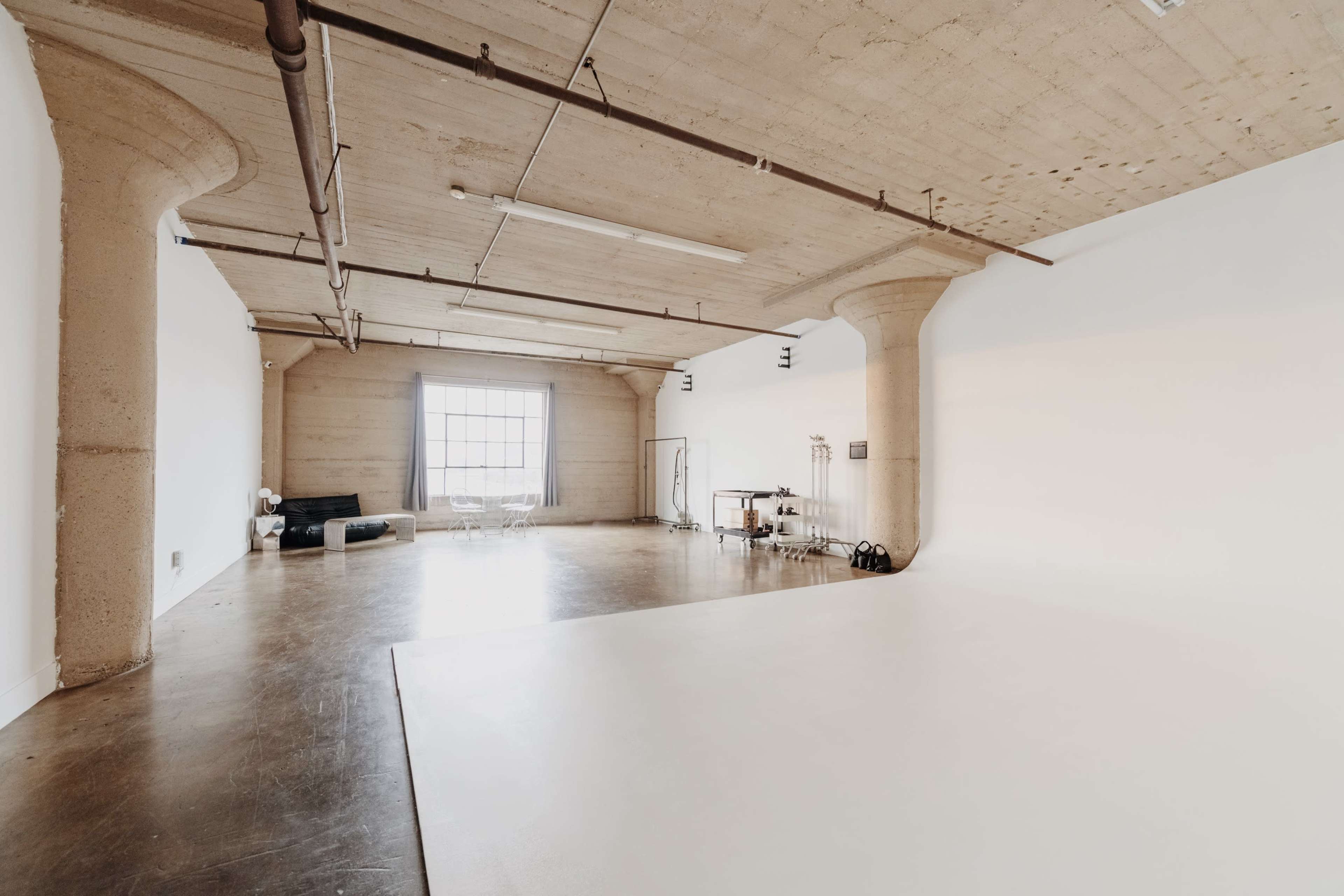 A spacious, minimalist studio with concrete walls, high ceilings, and a large window allowing natural light to enter.