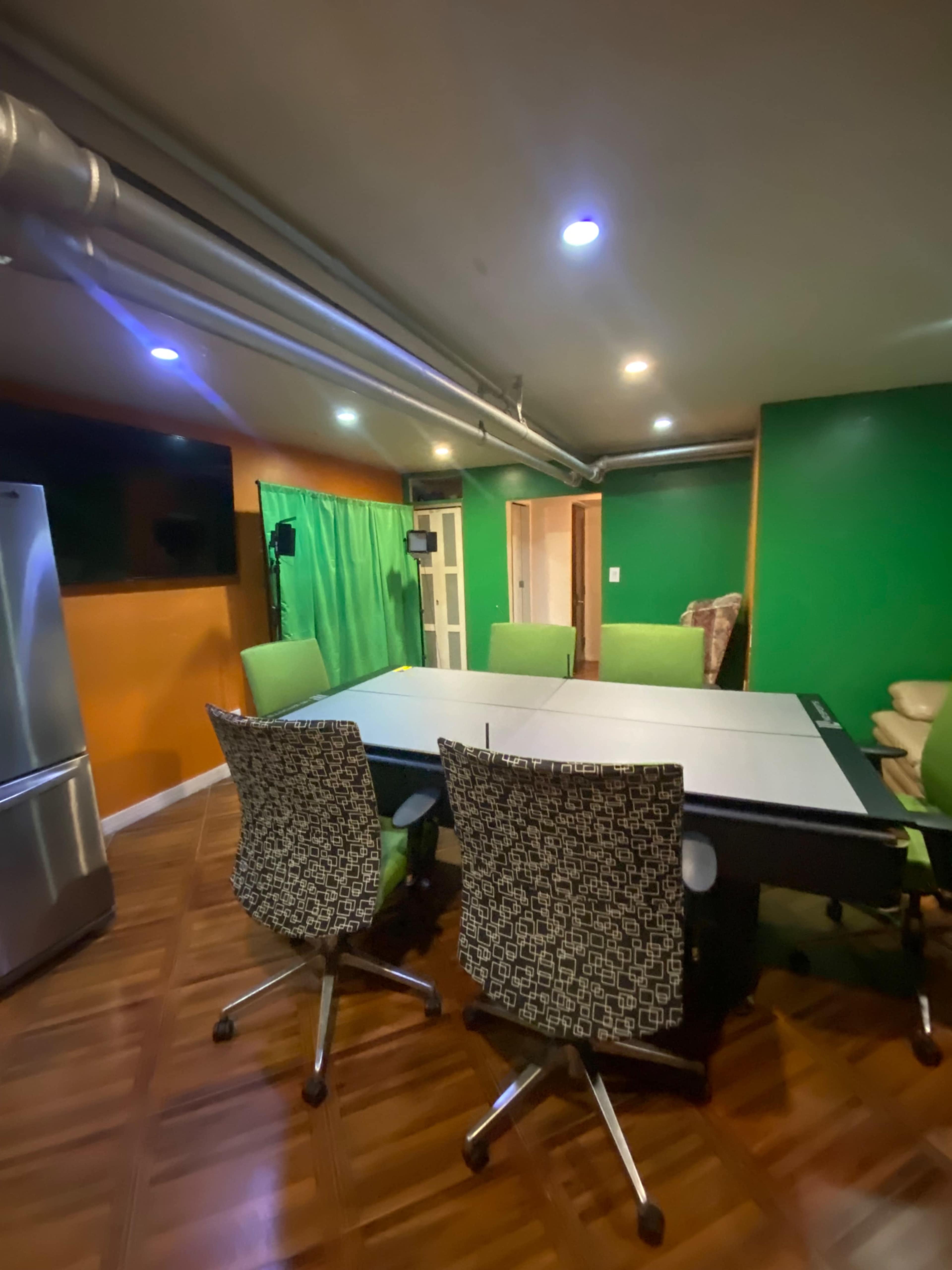 A modern conference room with a large table surrounded by ergonomic chairs, green walls, and a fridge in the corner.
