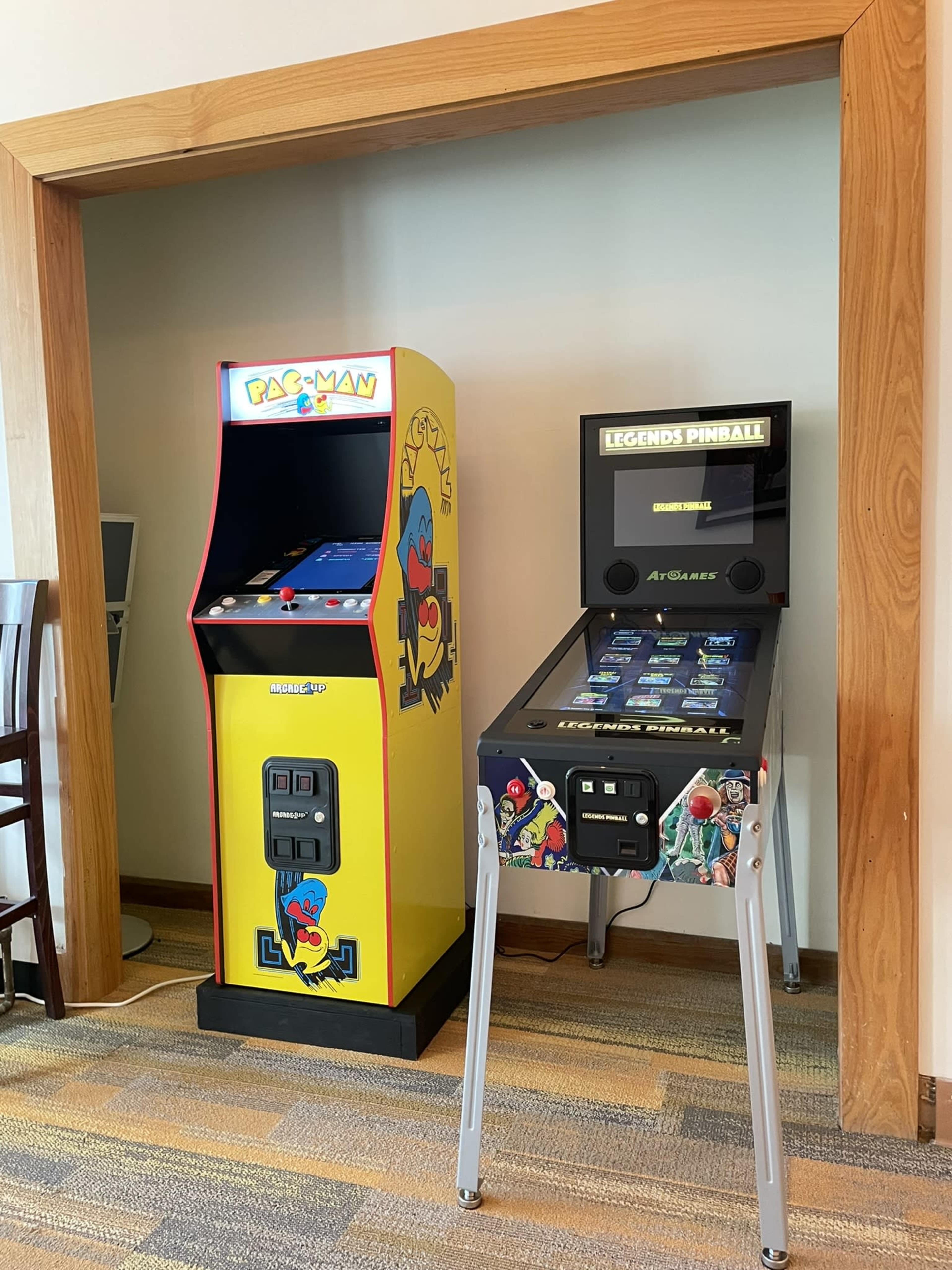 A classic Pac-Man arcade game machine beside a modern Legends Pinball arcade machine, both placed against a wall in a casual setting.