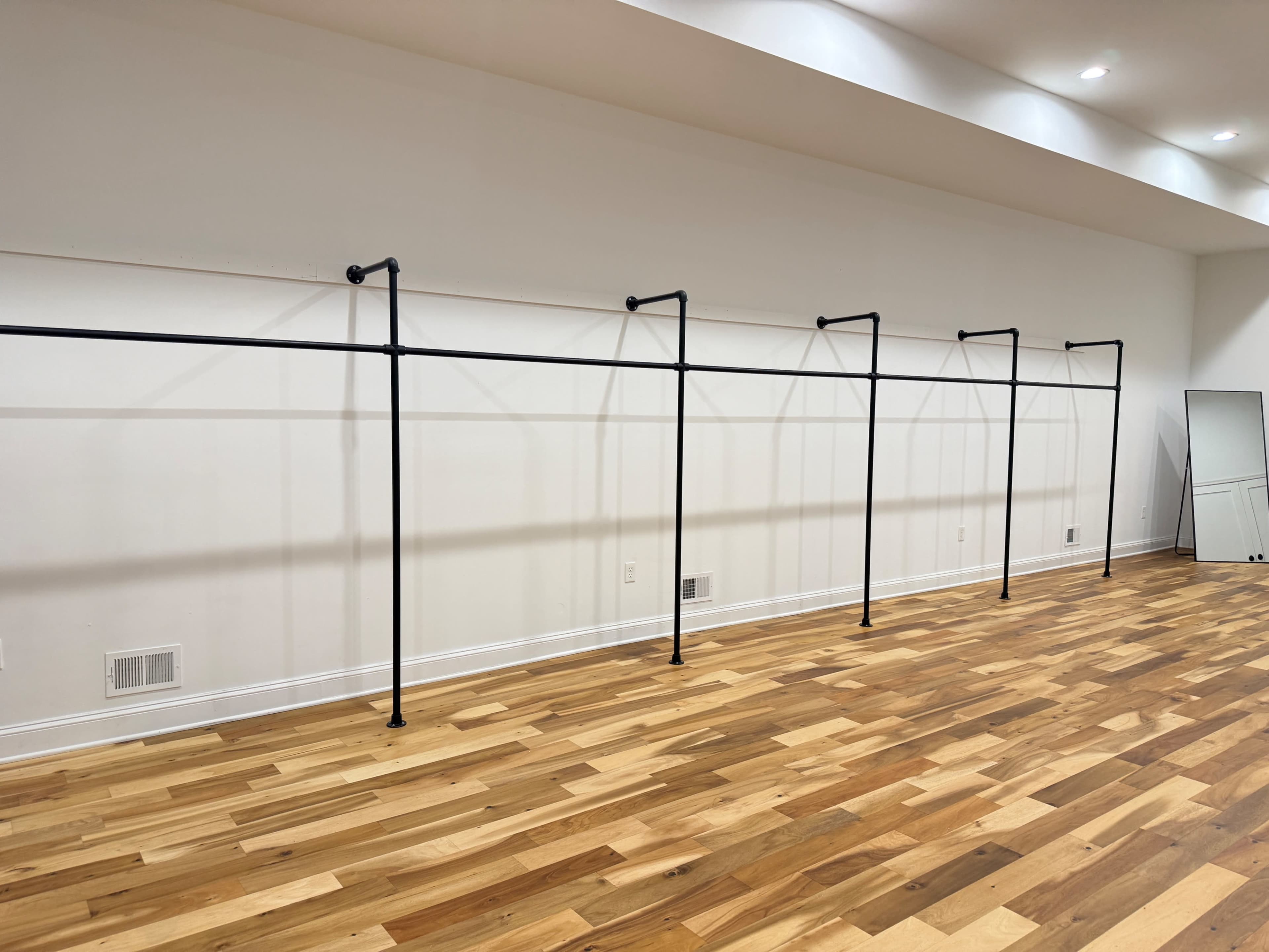 The image shows a row of black clothing racks against a white wall in a room with wooden flooring.
