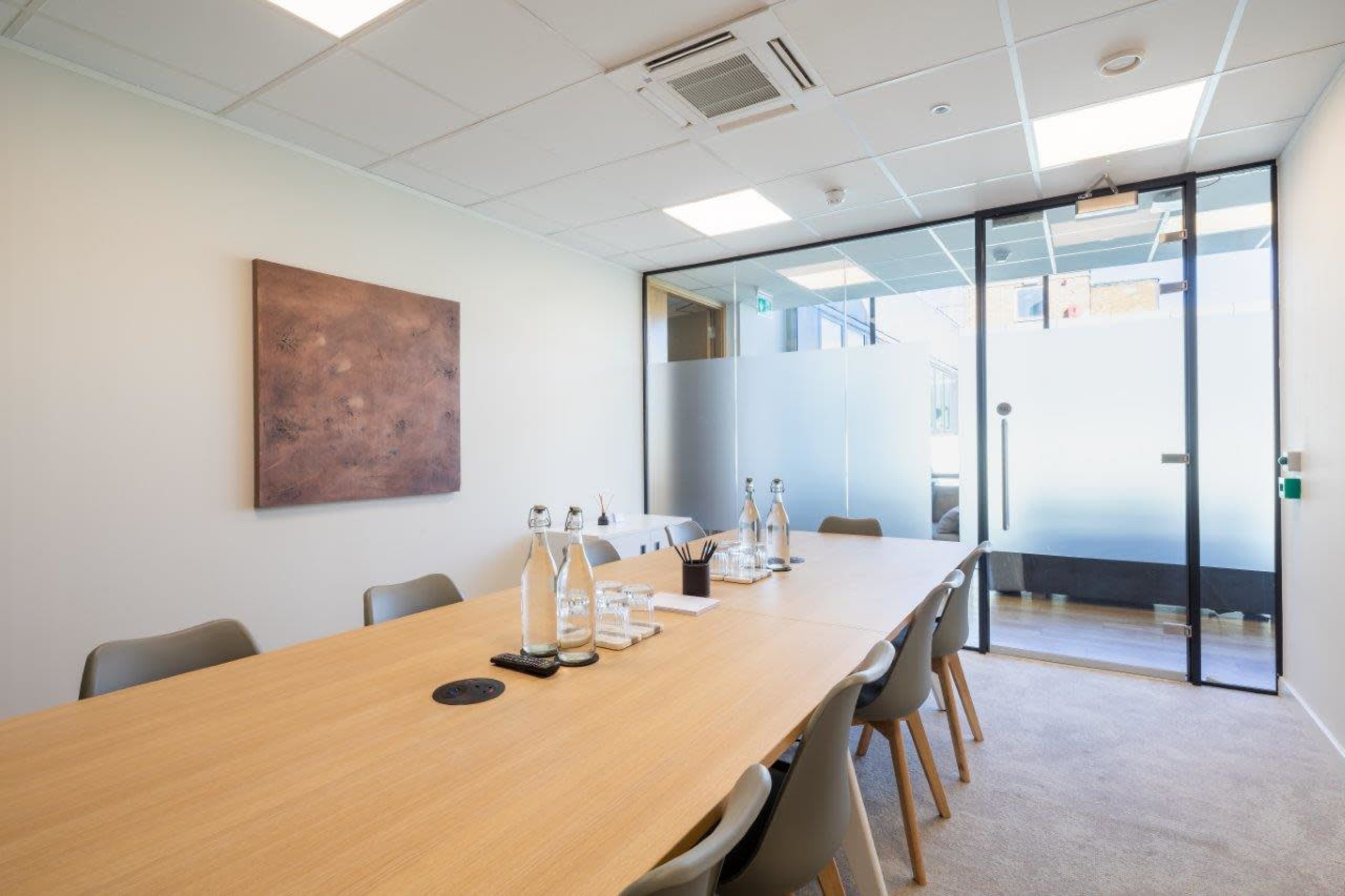 The image shows a modern conference room with a long wooden table, several water bottles, and a glass door leading to another room.