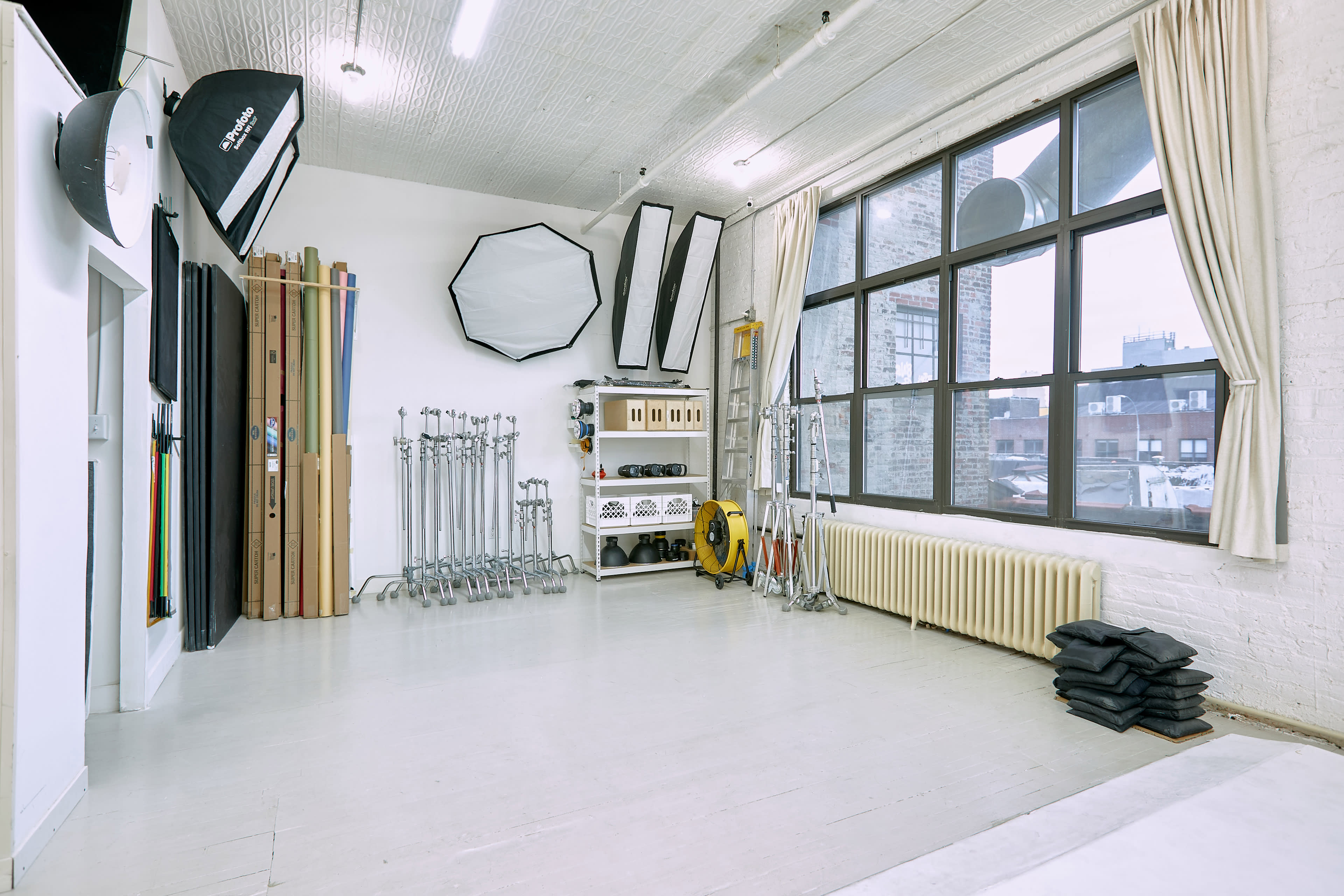 The image shows a well-equipped photography studio featuring various lighting equipment, backdrops, and a large window providing natural light.