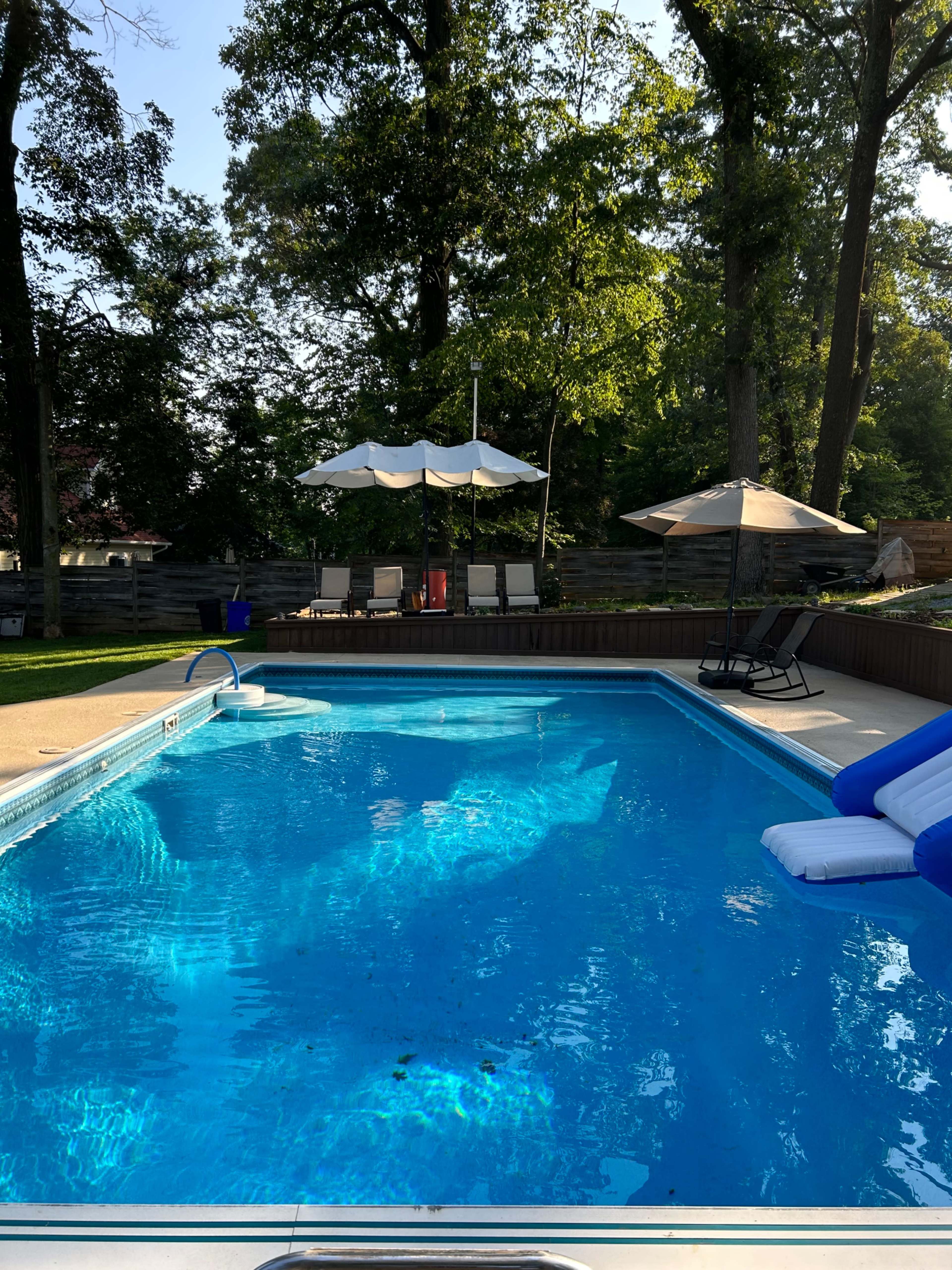 A clear swimming pool surrounded by trees, with lounge chairs and an umbrella in the background.