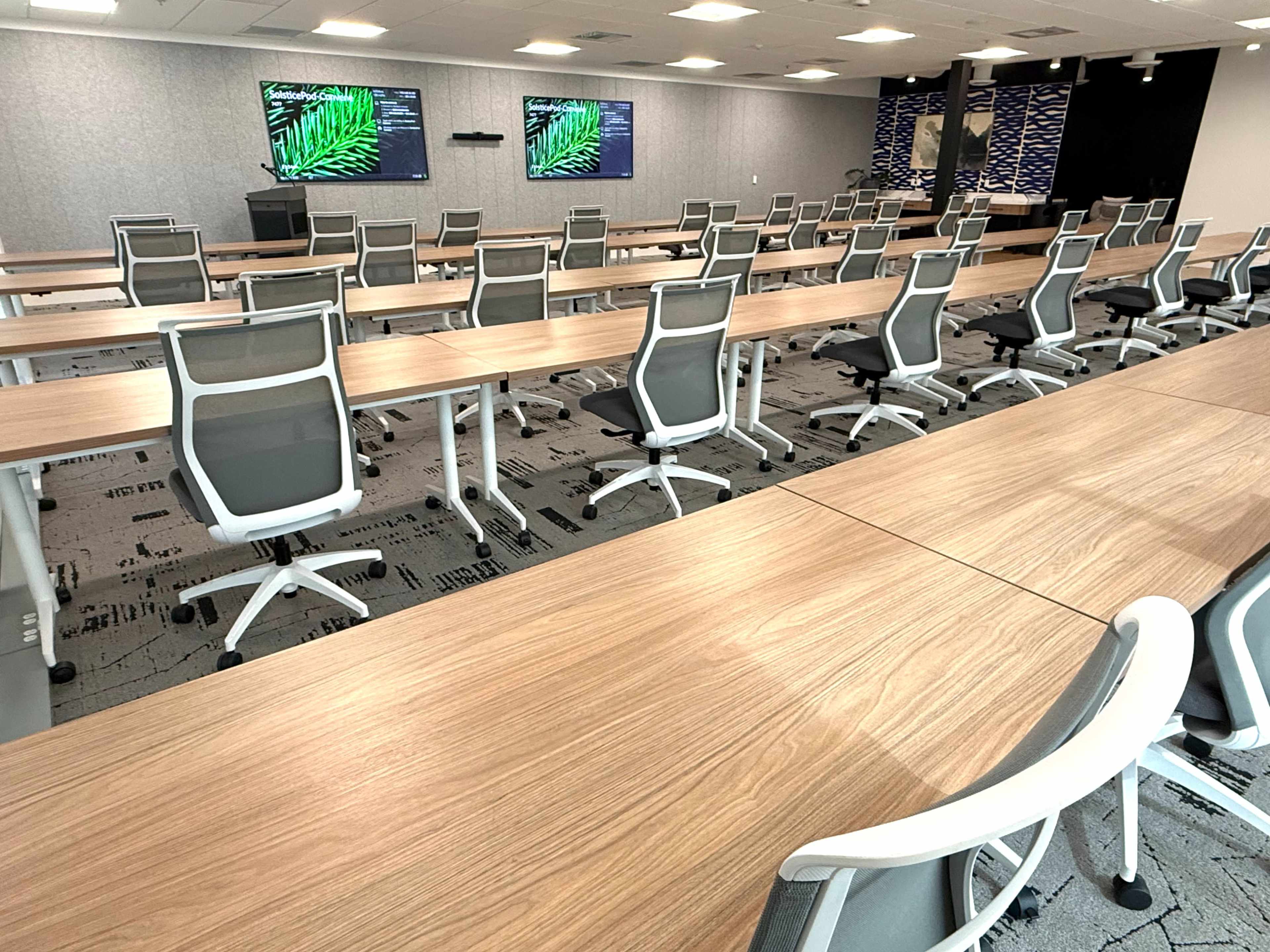 A modern conference room features multiple long tables arranged in a U-shape with ergonomic chairs and two screens displaying tropical images.