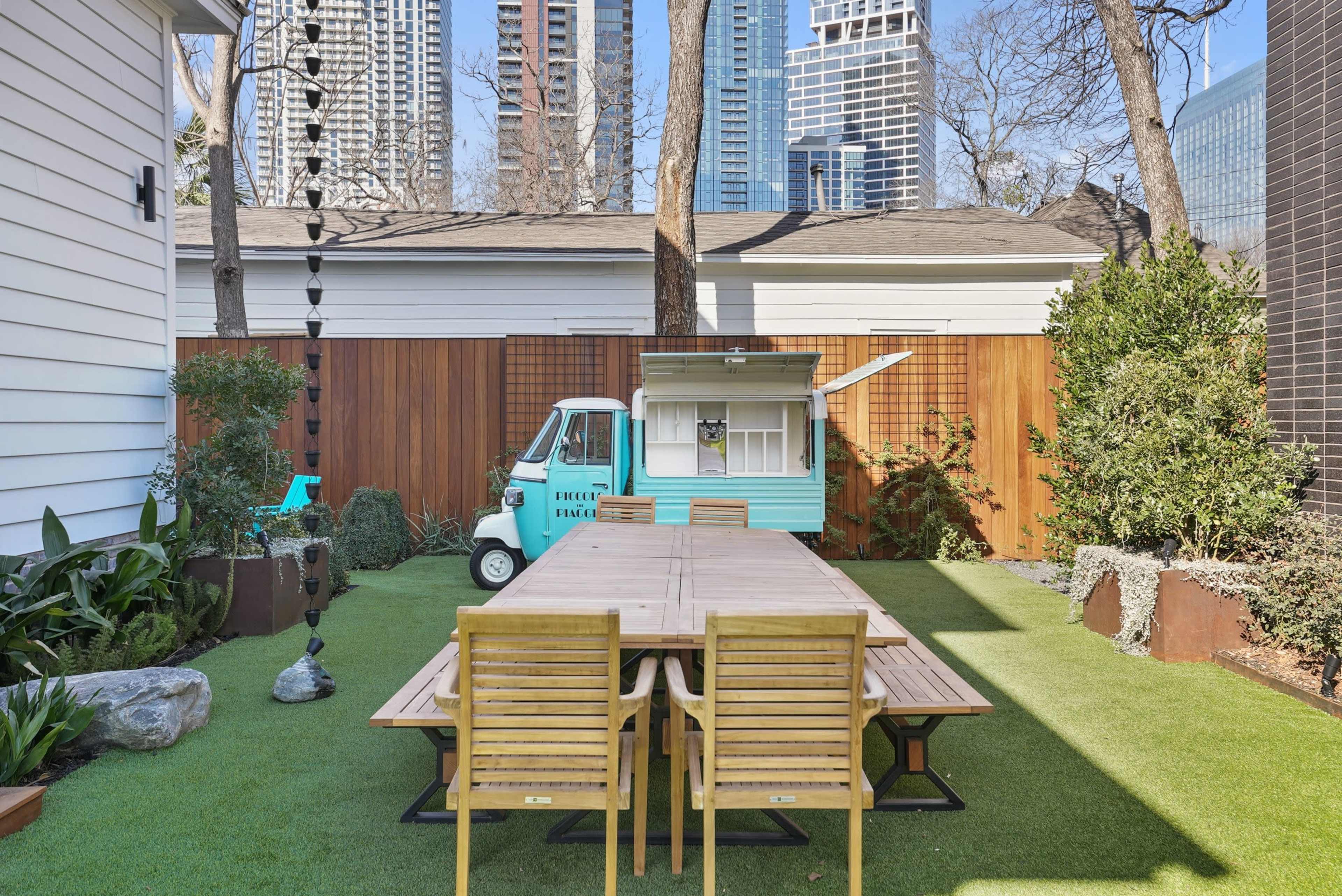 A modern outdoor space features a large wooden dining table surrounded by chairs, with a vibrant food truck and tall city buildings in the background.
