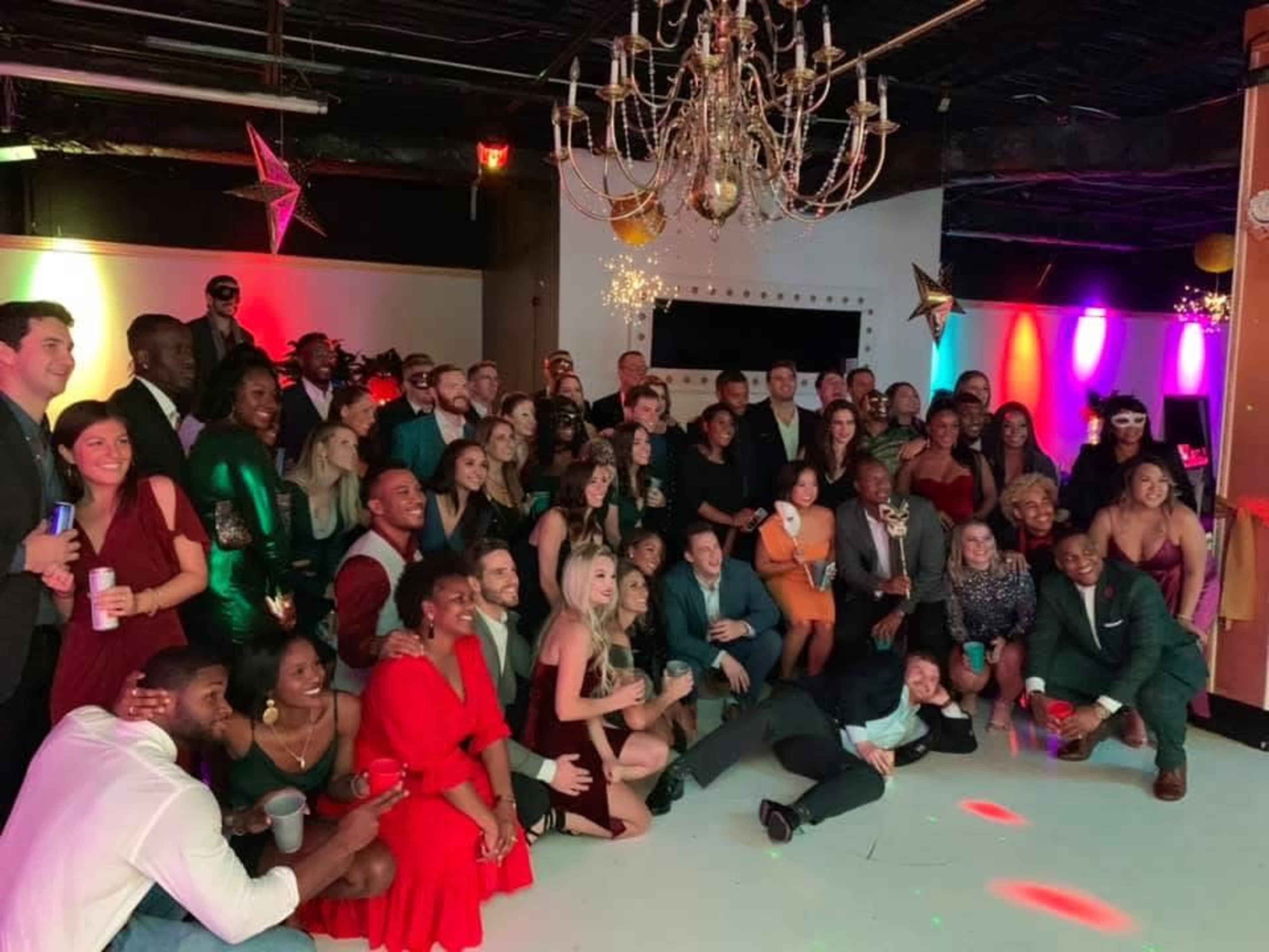 A large group of people is gathered in a decorated venue, posing for a photo under a chandelier with colorful lights in the background.