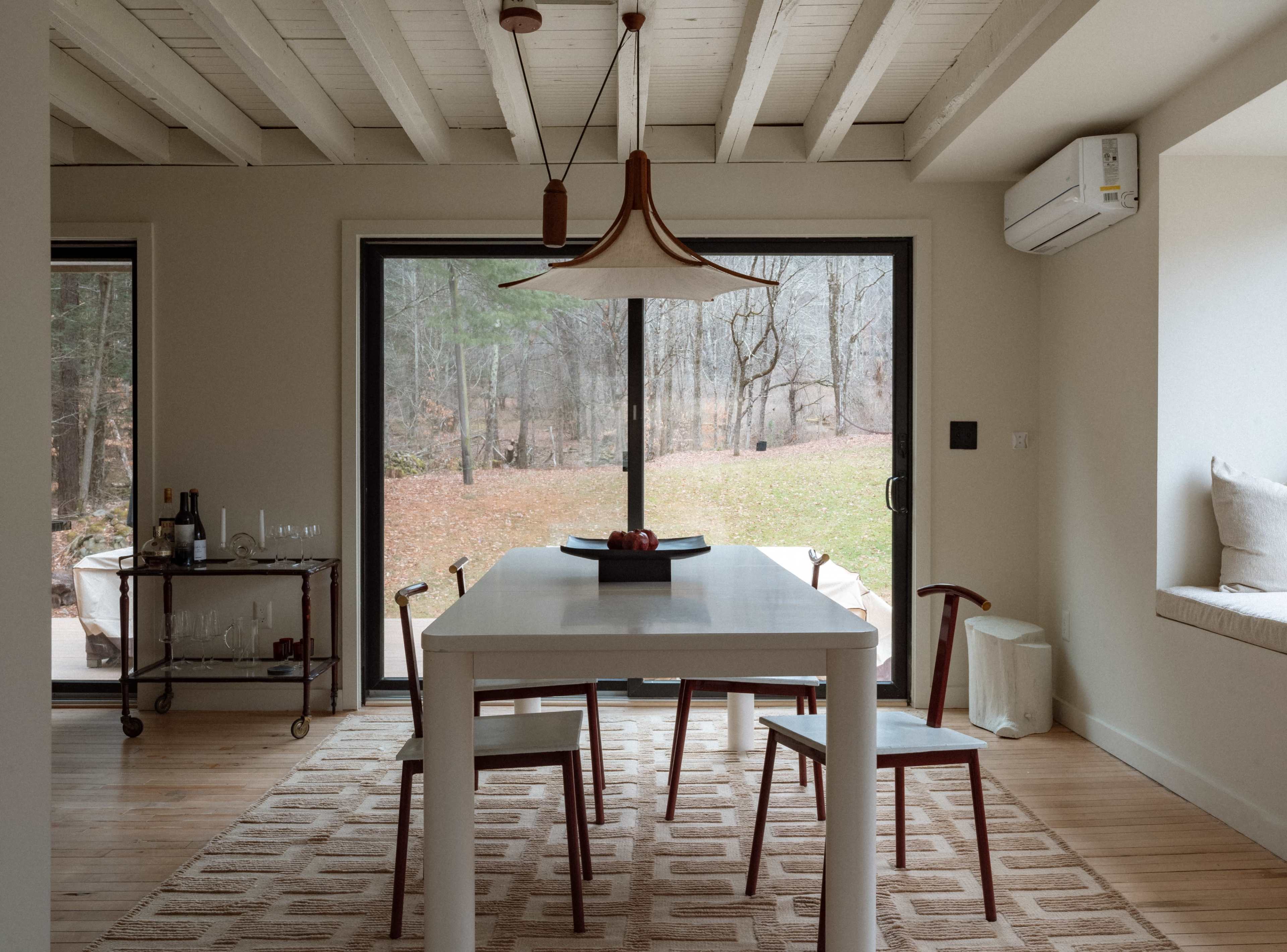 A minimalist dining area features a white table surrounded by four chairs, with large glass doors leading to a wooded outdoor view.