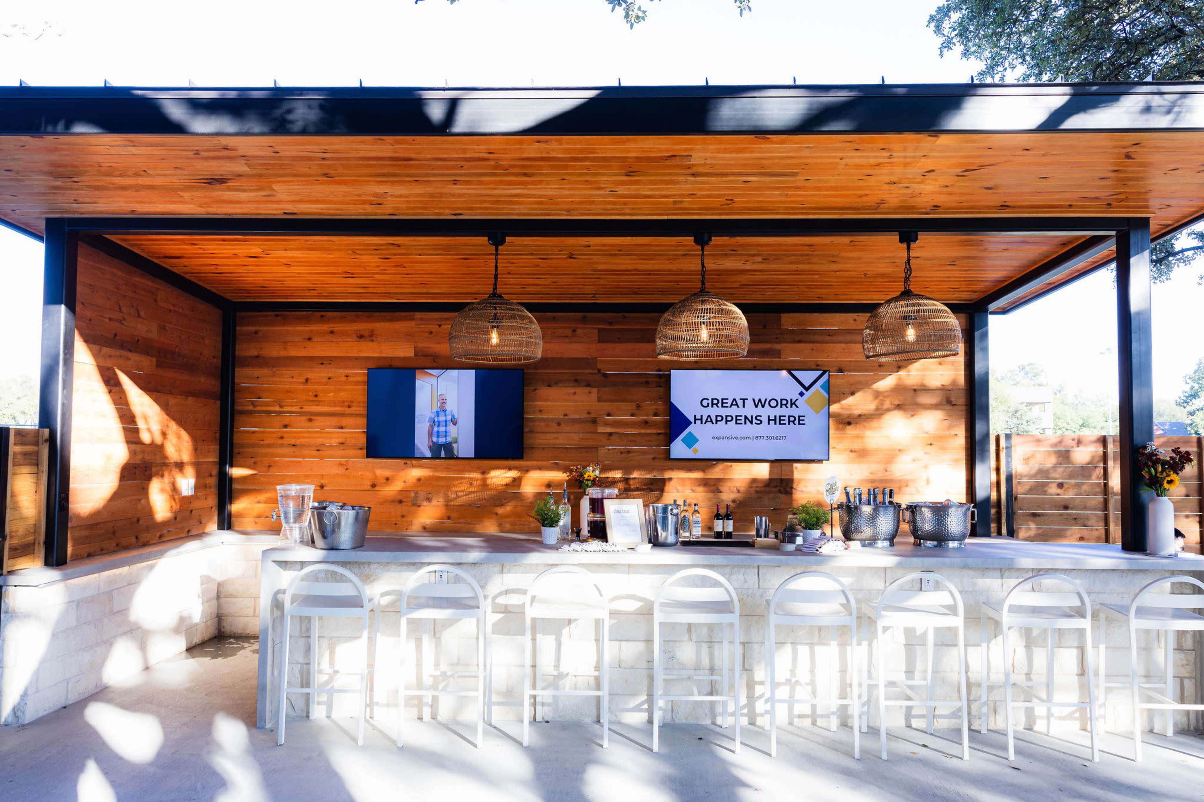 The image shows an outdoor bar area with a wooden ceiling, stone countertop, and decorative pendant lights, featuring a large screen displaying a presentation.