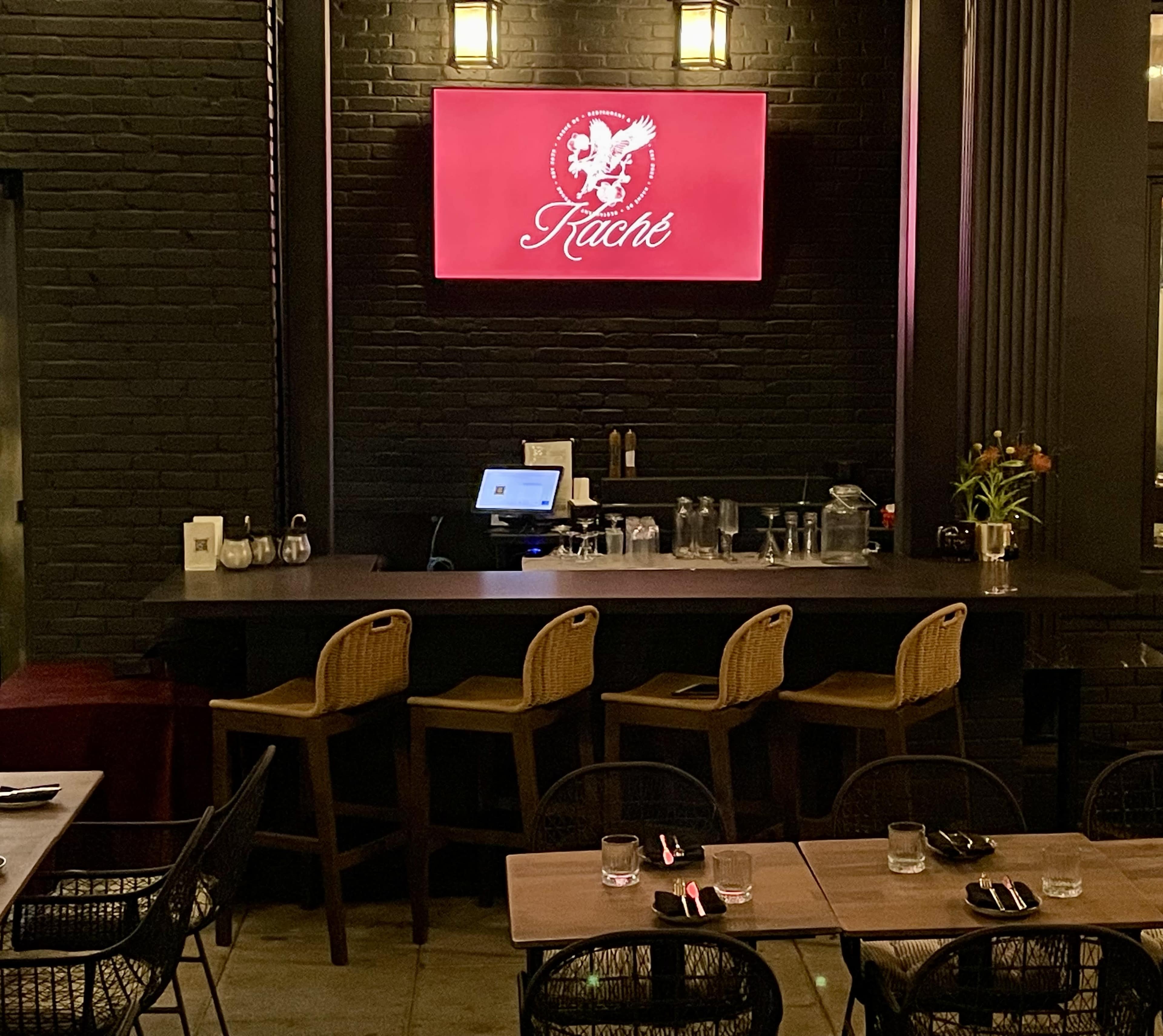 The image shows a restaurant interior featuring a bar with tall stools, a digital screen displaying the restaurant's name, and a dark brick wall.