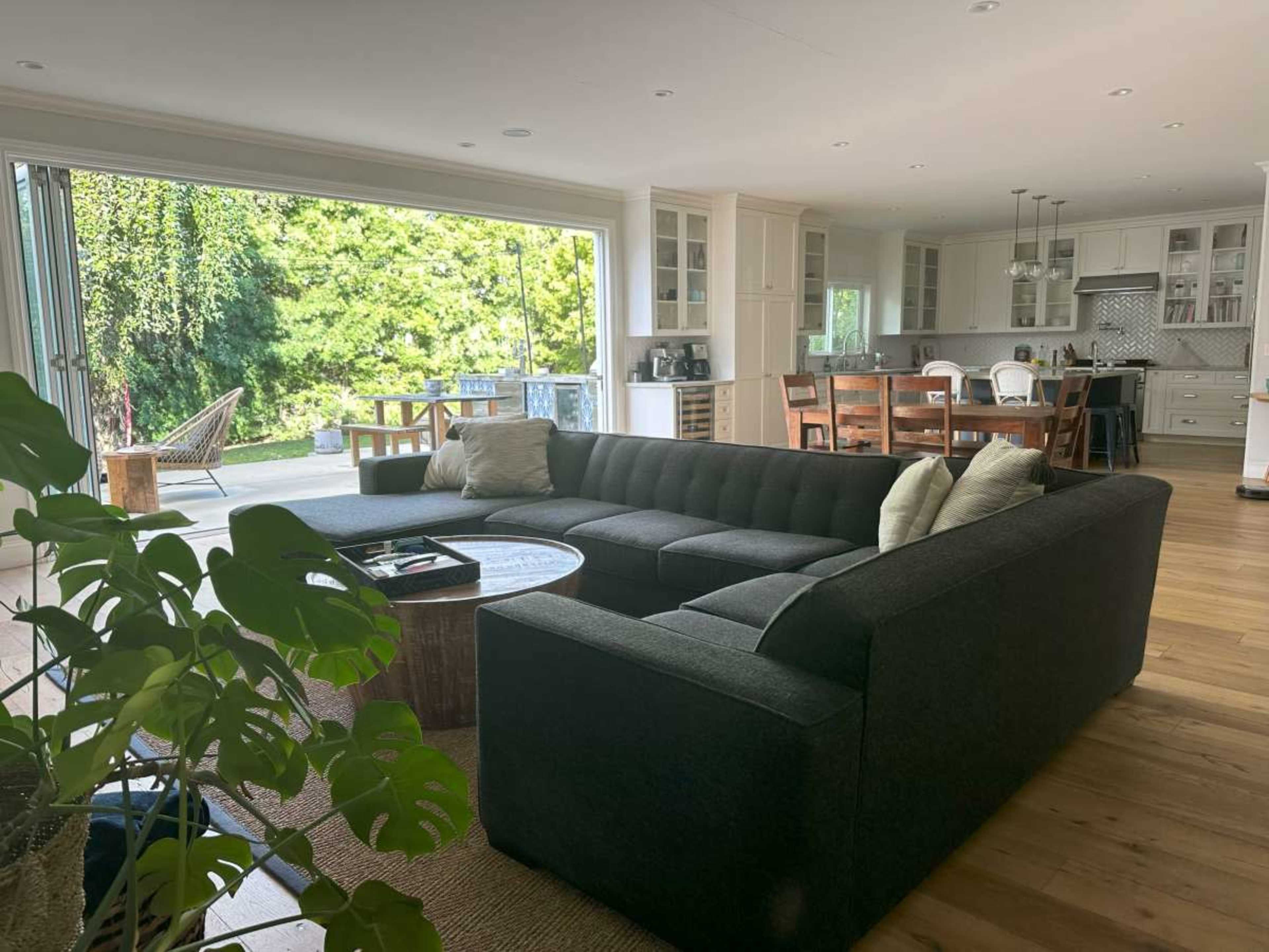 A spacious living area features a dark sectional sofa, a round coffee table, and large glass doors leading to an outdoor patio with greenery.