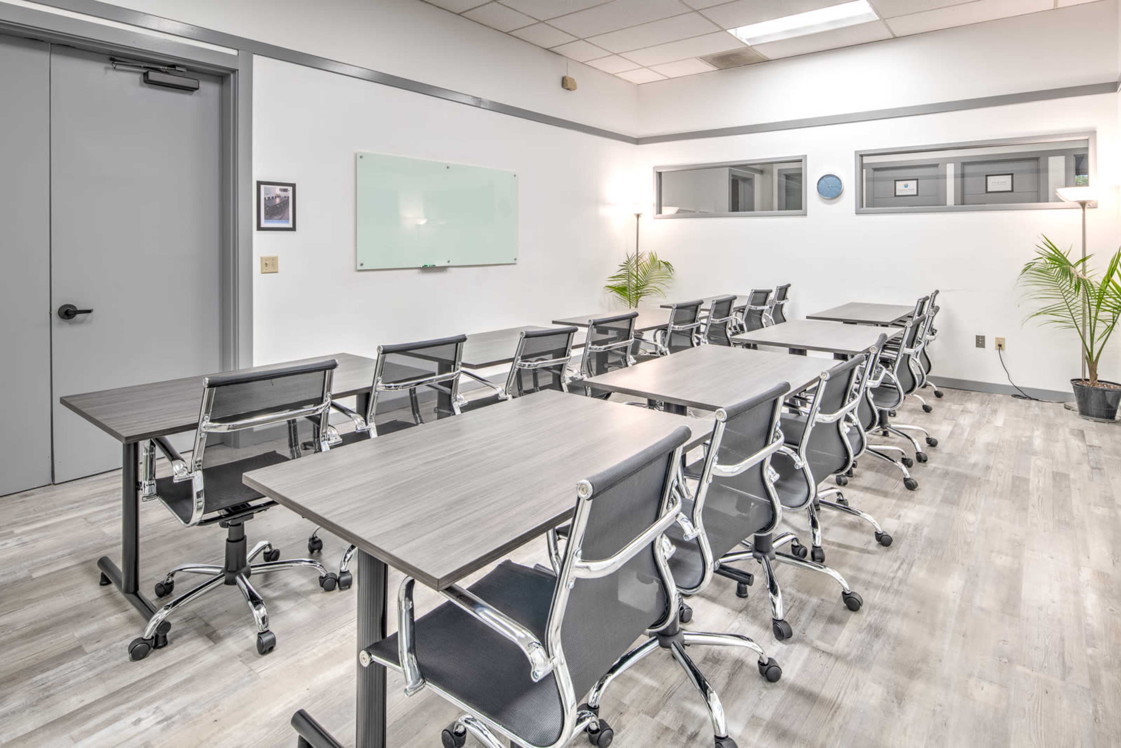 A modern conference room with several rows of tables and chairs, equipped with a whiteboard and plants.
