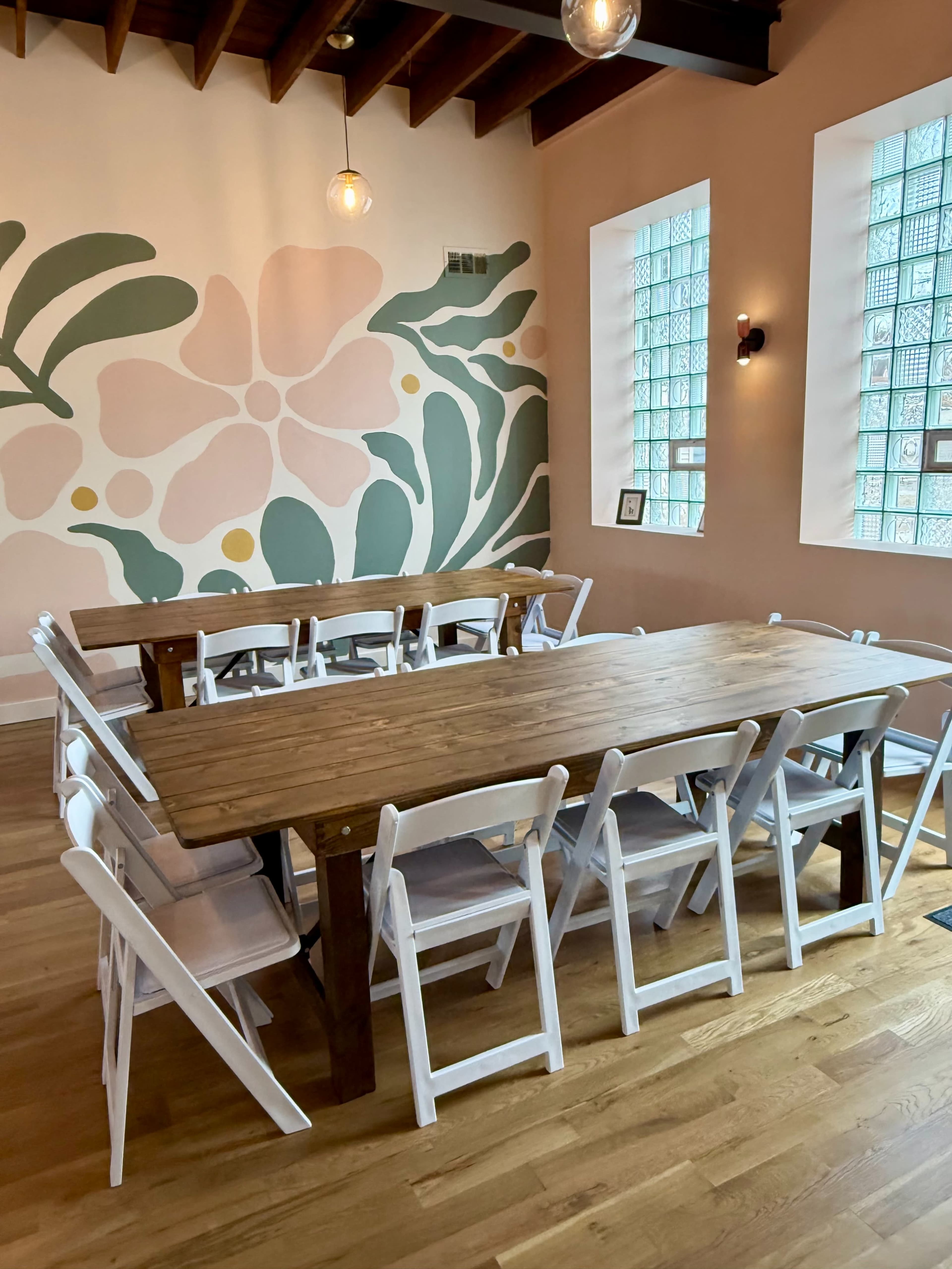 A long wooden table surrounded by white folding chairs is placed in a brightly lit room with a floral mural on one wall.