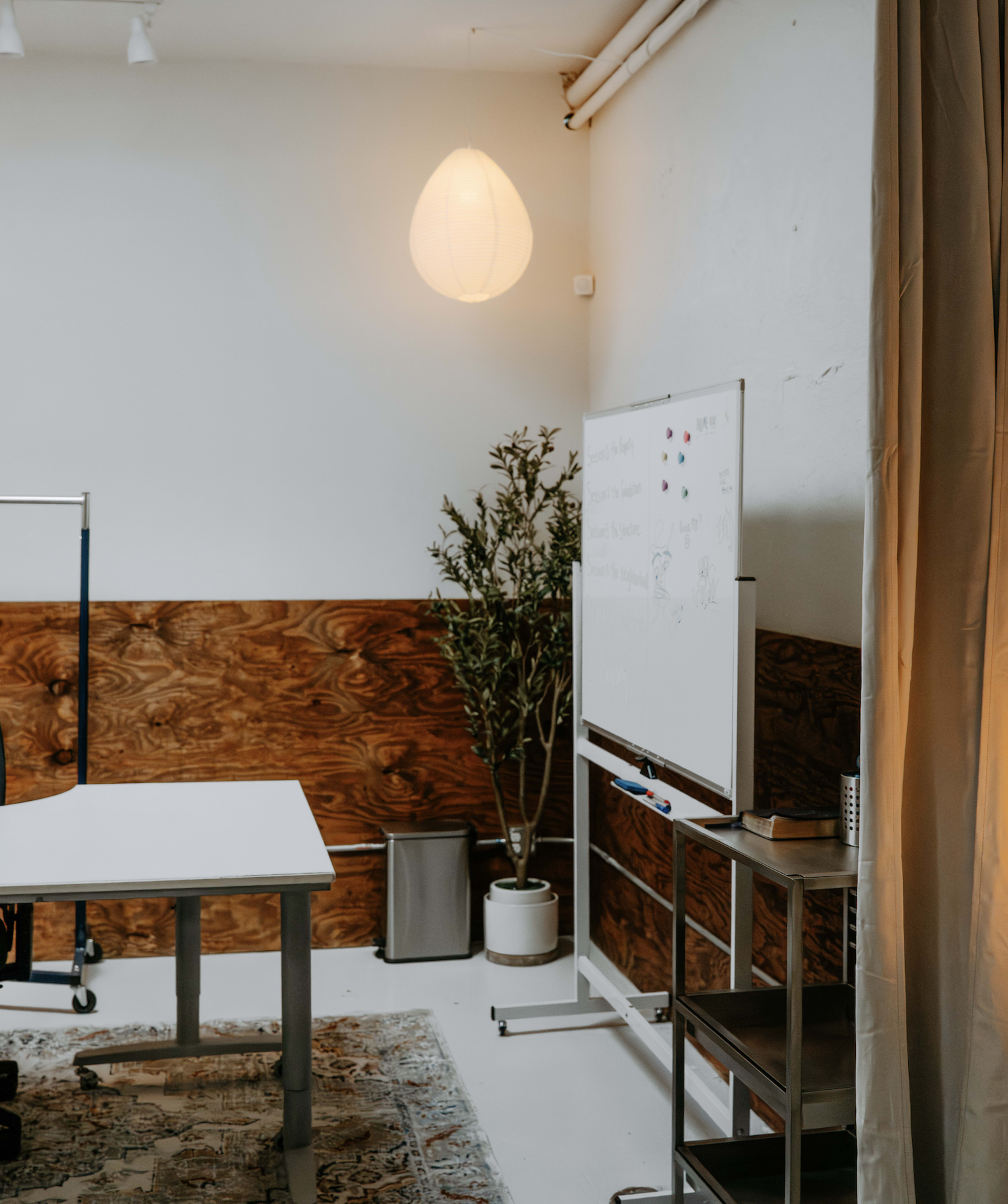 The image shows a minimalist office space with a whiteboard, a plant, and a desk situated against a wooden wall.