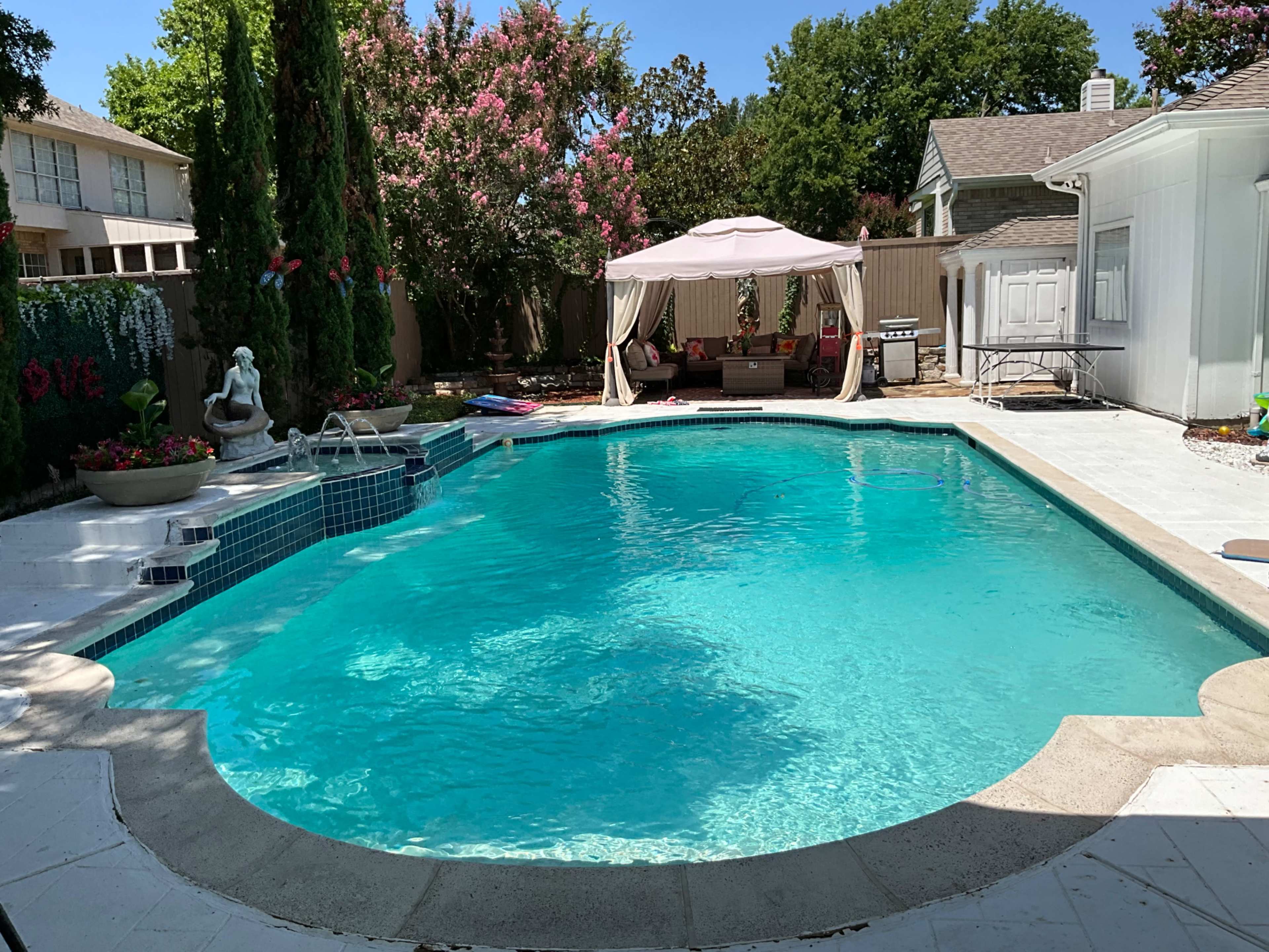 The image shows a well-maintained swimming pool surrounded by greenery, with a shaded seating area off to one side.