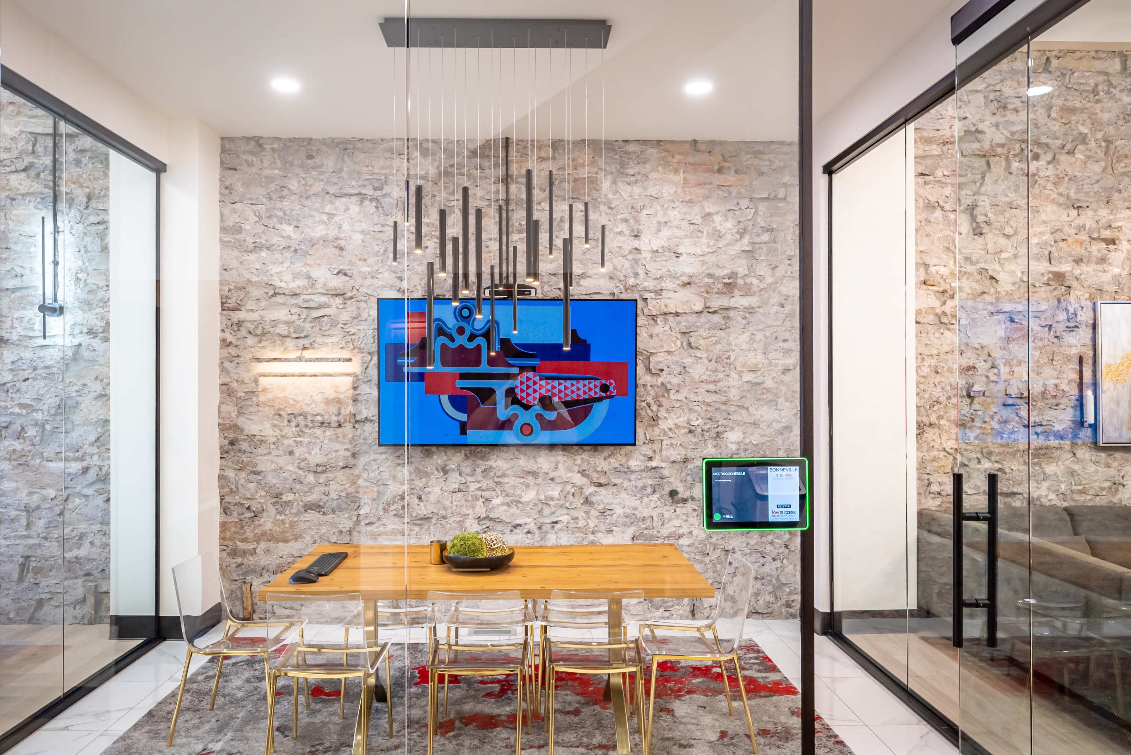 A modern dining area features a long wooden table surrounded by clear acrylic chairs, with a stone accent wall and a large screen displaying abstract art.