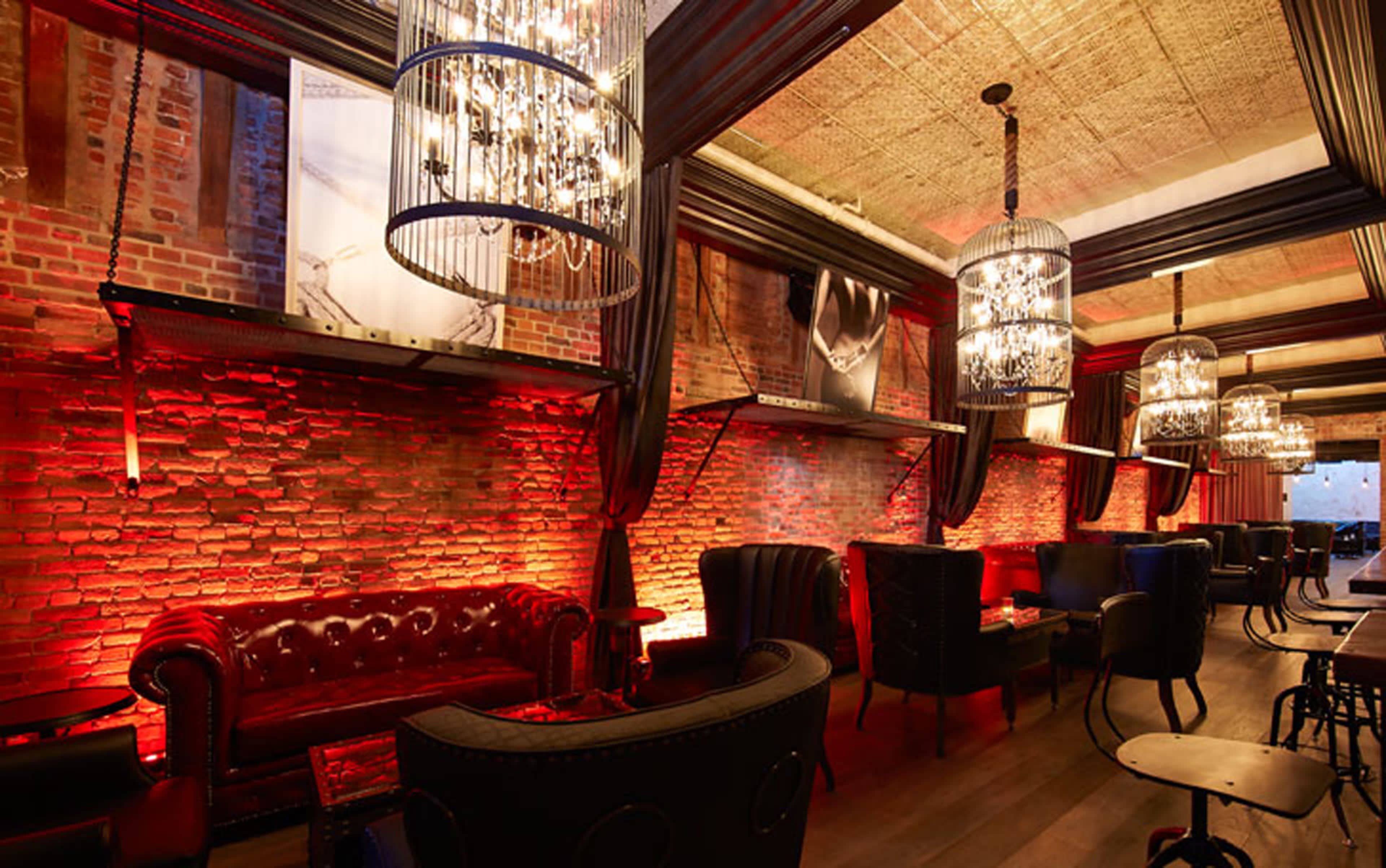 The image shows a dimly lit lounge featuring exposed brick walls, ornate chandeliers, and plush seating arrangements.