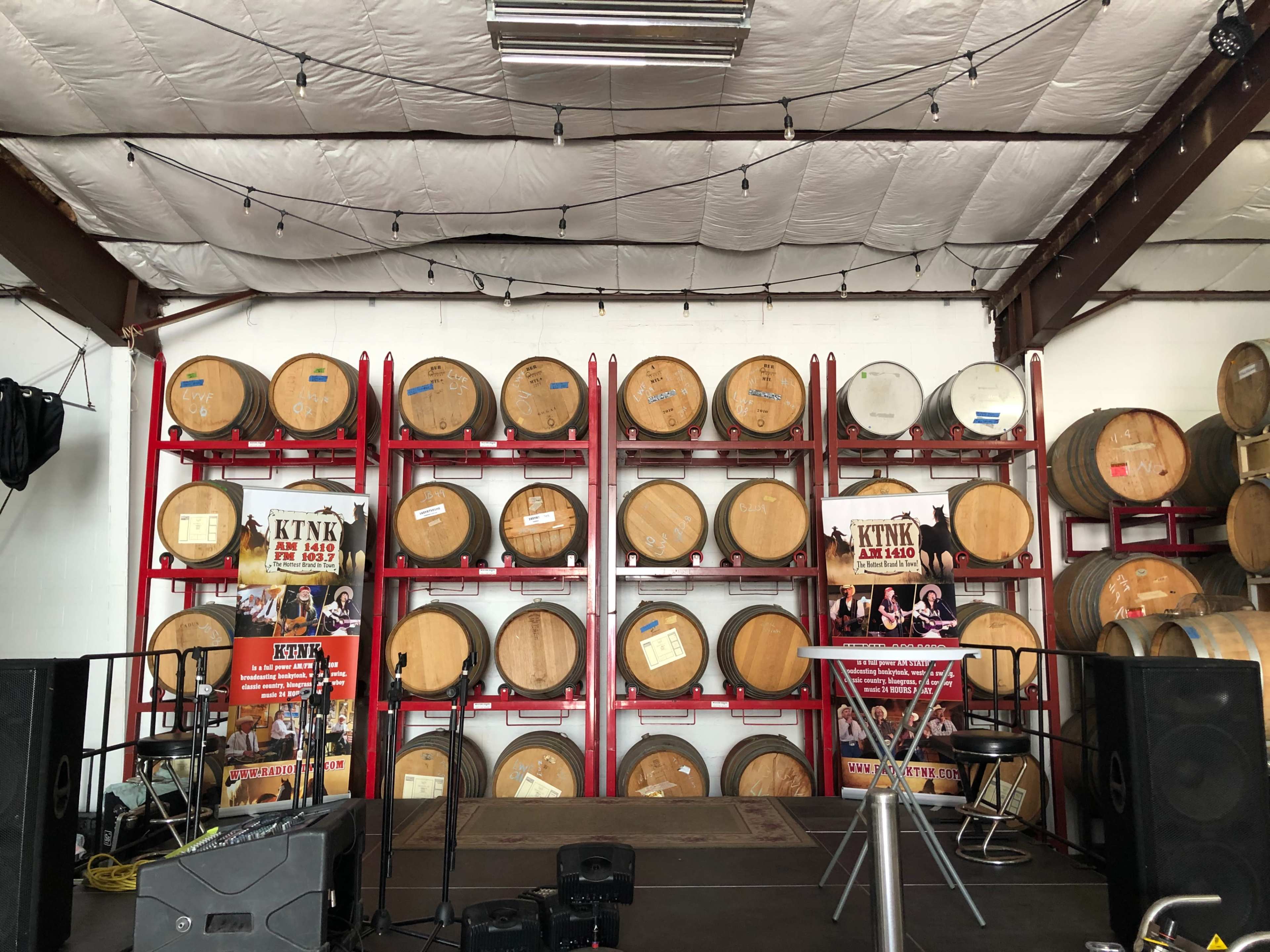 A staged area with wooden barrels stacked on red metal racks in a warehouse setting, accompanied by promotional posters.