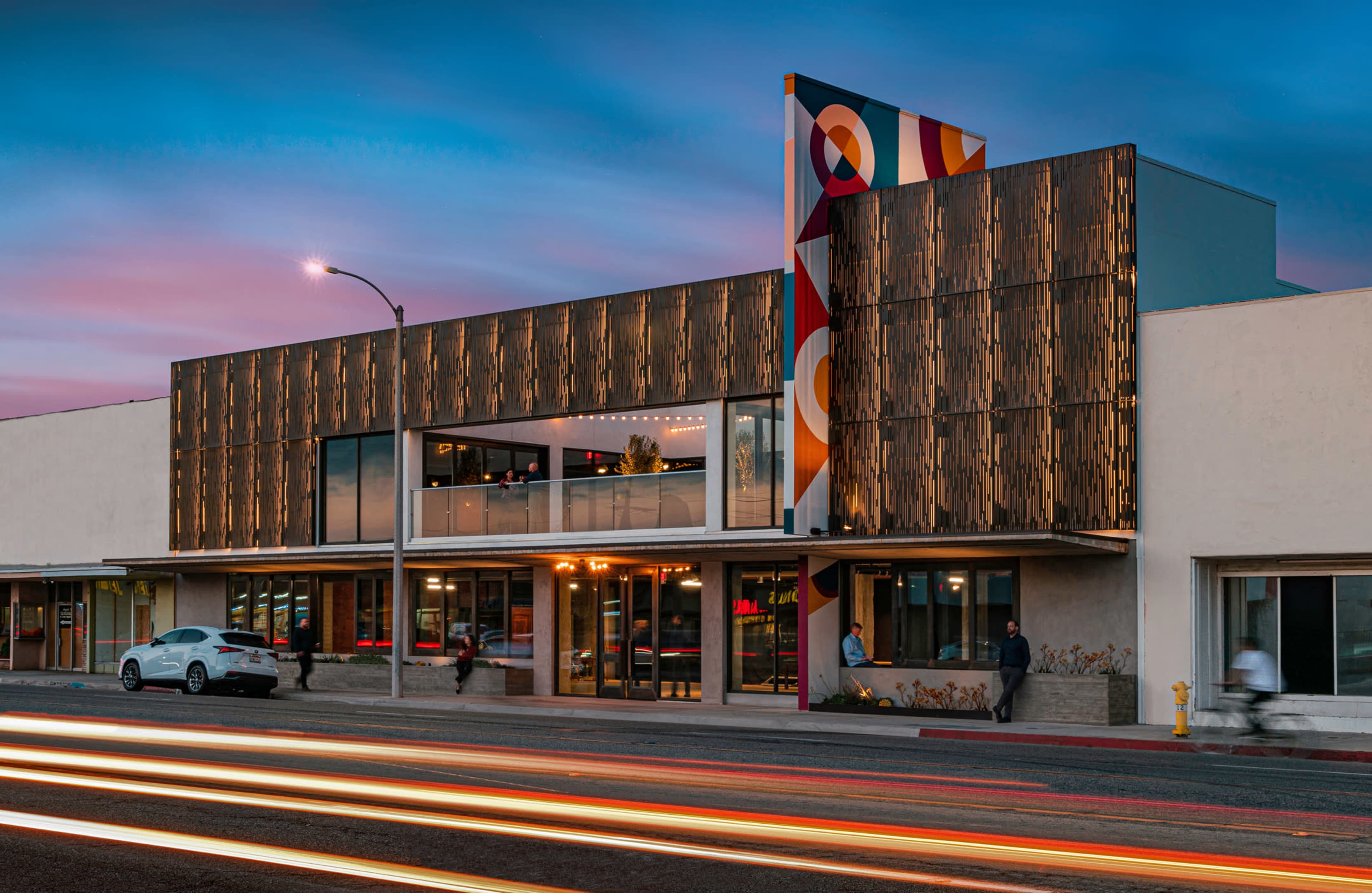 The image shows a modern building facade at dusk, featuring a colorful artistic mural and illuminated interior spaces, with streaks of light from passing cars in the foreground.