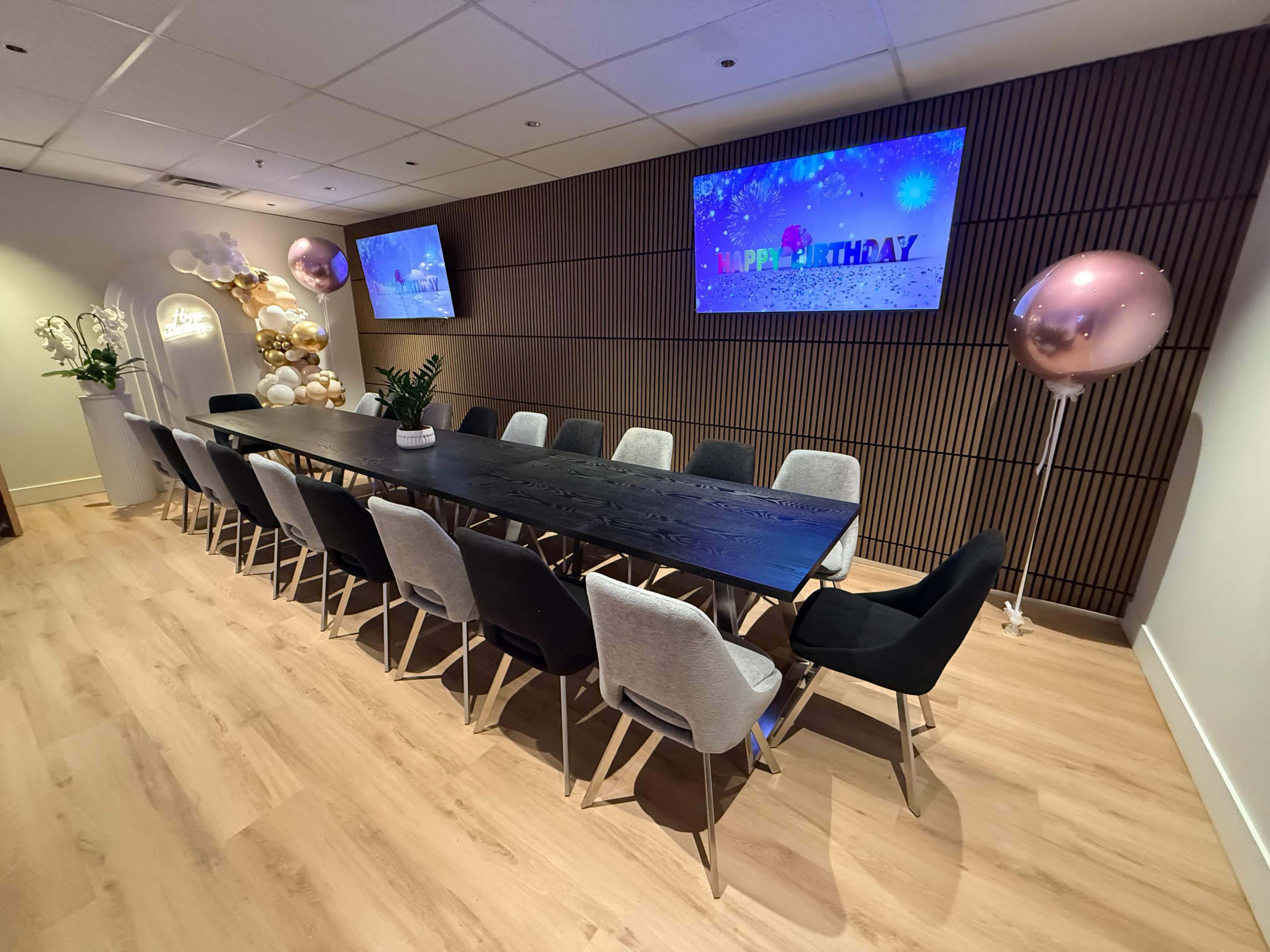 A long black conference table with chairs is set up in a decorated meeting room featuring screens displaying a "Happy Birthday" message and balloons.