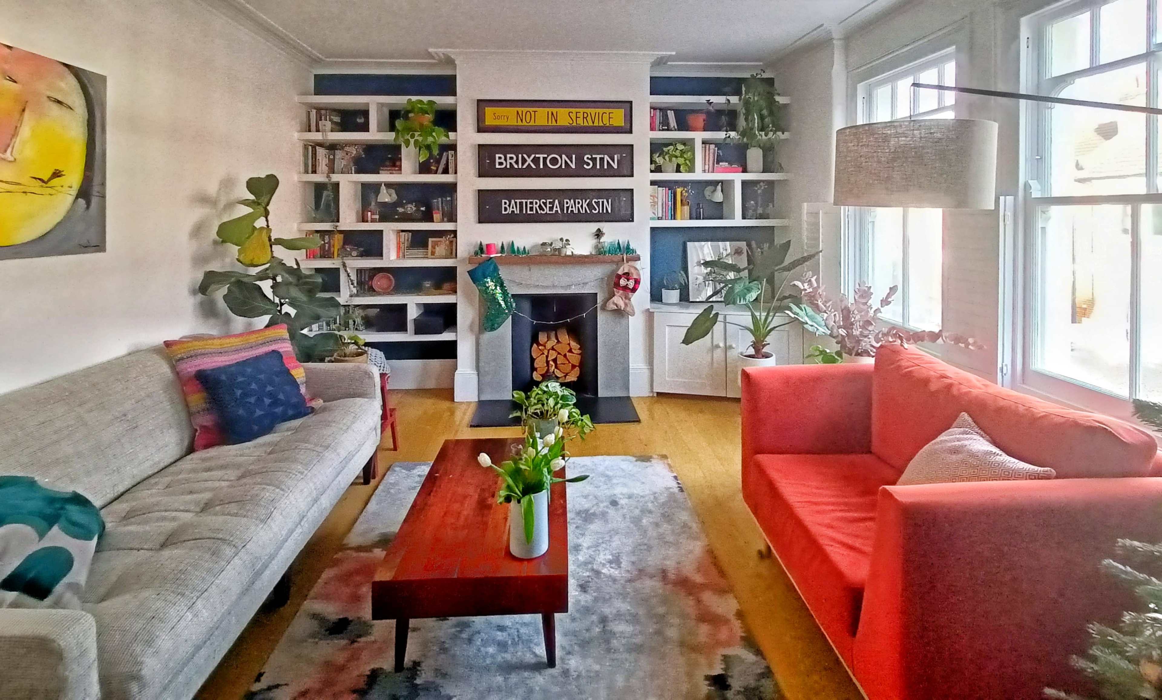 The living room features a gray sofa, a bright red couch, a wooden coffee table, and shelves lined with books and decorations, with a fireplace at its center.